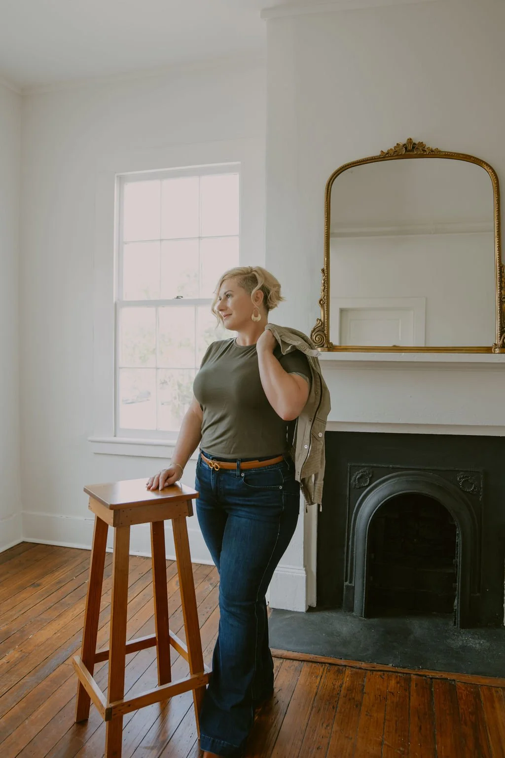 A woman with short blond hair, wearing a green T-shirt and jeans, stands in a bright room with a wooden floor, next to a small wooden table, near a window with white frames, and a fireplace with a decorative mirror above it.