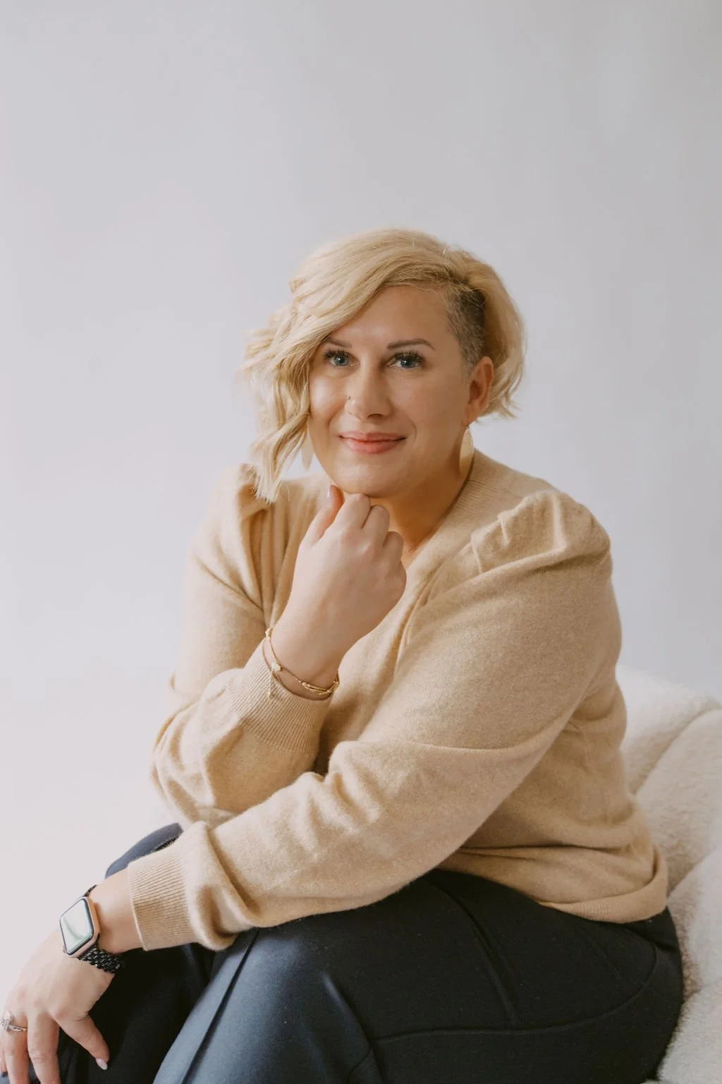 A woman with blonde, wavy hair and fair skin, sitting on a beige couch with a white background, smiling, wearing a beige sweater, black pants, a gold bracelet, and a smartwatch.