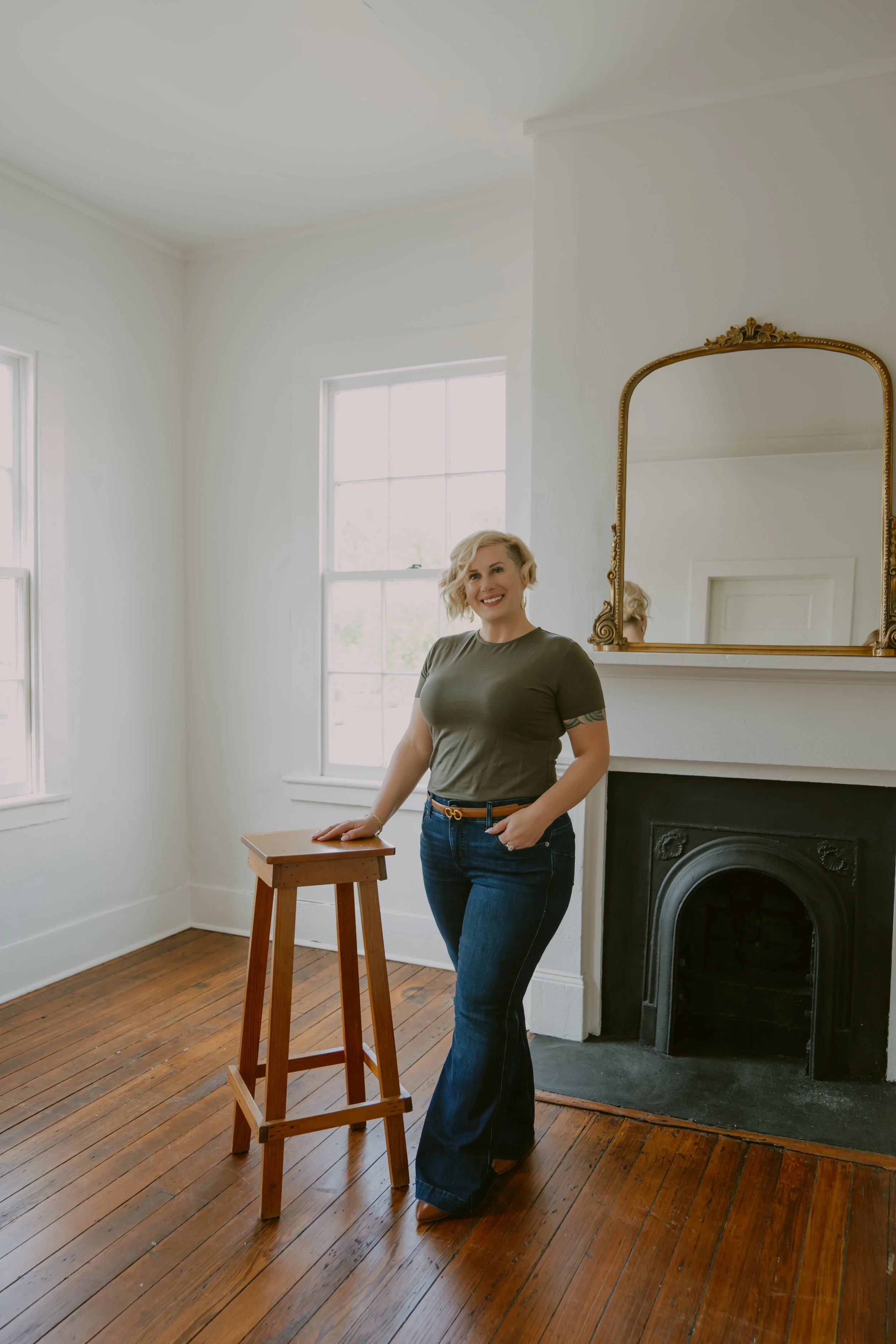 A woman with blonde hair standing in a bright room with white walls, wooden floors, and large windows. She's smiling and leaning on a small wooden stool next to a fireplace with a gold-framed mirror above it.