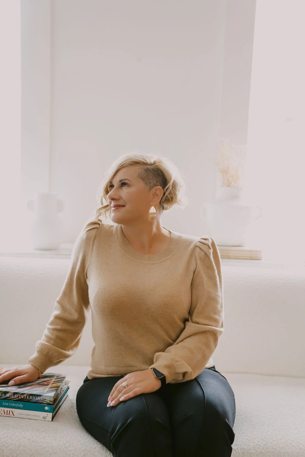 A woman with short blonde hair sitting on a cream-colored couch, looking to her left with a gentle smile. She is wearing a beige sweater and dark pants, with a smartwatch on her wrist. There are magazines and a stack of books on her lap, and a white wall with decorative vases in the background.