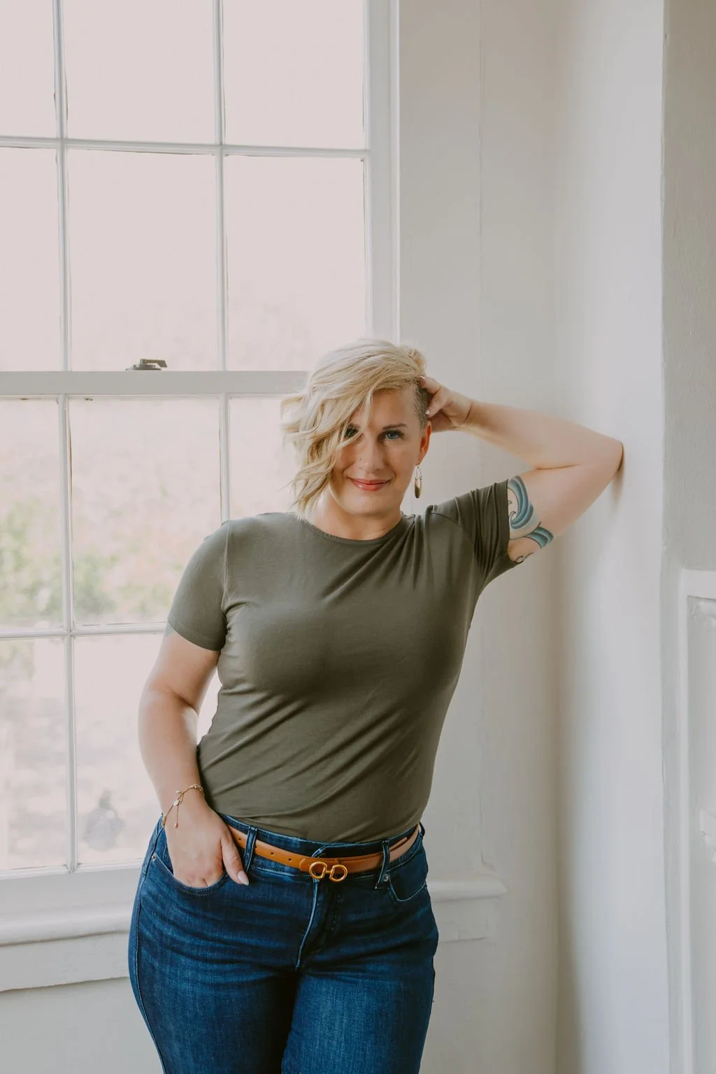 A woman with short blonde hair and a tattoo on her arm standing by a window, wearing a green t-shirt and blue jeans.