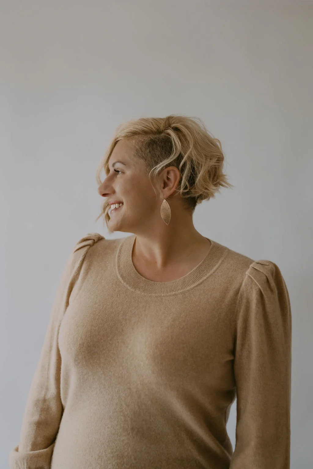 A woman with short, wavy blonde hair, wearing a beige sweater and large, gold, oval earrings, smiling and looking to the left against a plain light background.