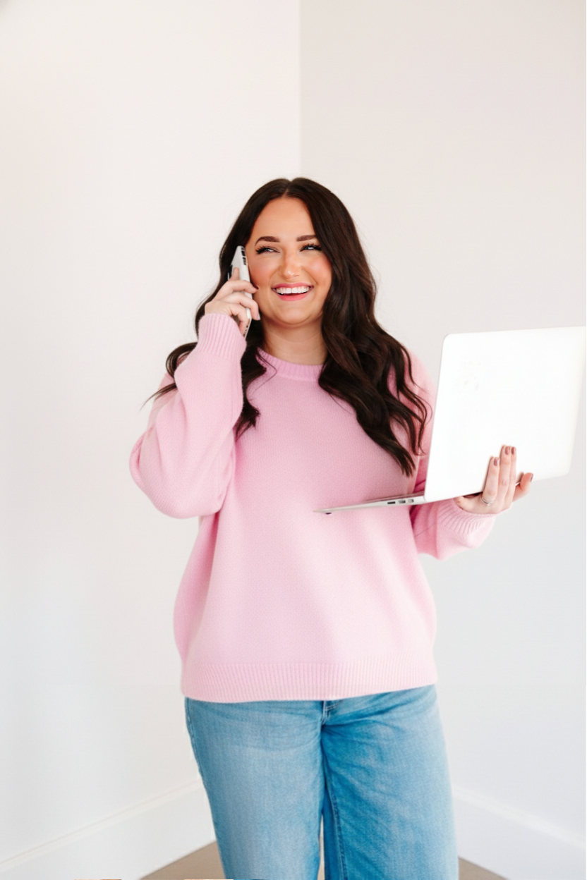 Young woman in pink sweater holding a laptop and talking on a cell phone, smiling.