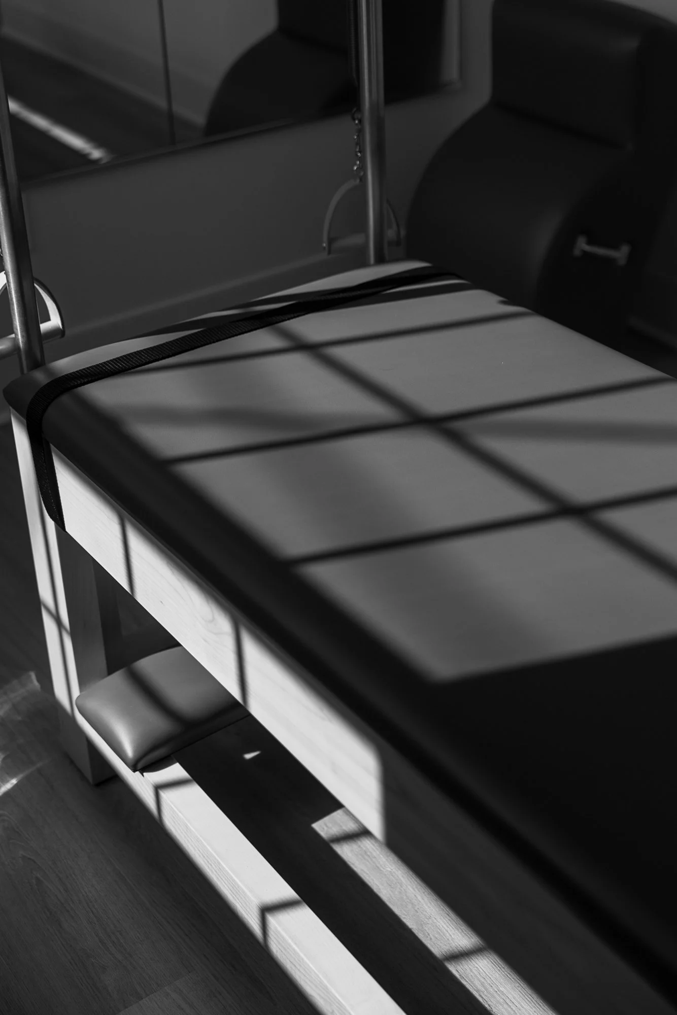 A black-and-white photo of a modern table with a glass top, shadowed by natural light through a window, with a chair and part of a kitchen visible in the background.