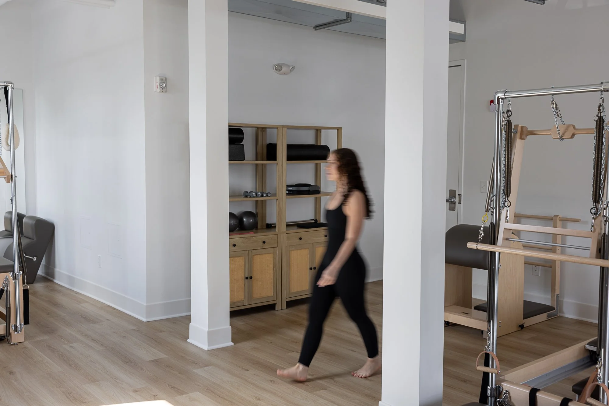 A woman in black workout clothes walking through a space with fitness equipment, including a wooden Pilates reformer, a shelf with black exercise balls and foam rollers, and part of a Pilates Cadillac machine, in a bright, white-walled fitness studio with wood flooring.