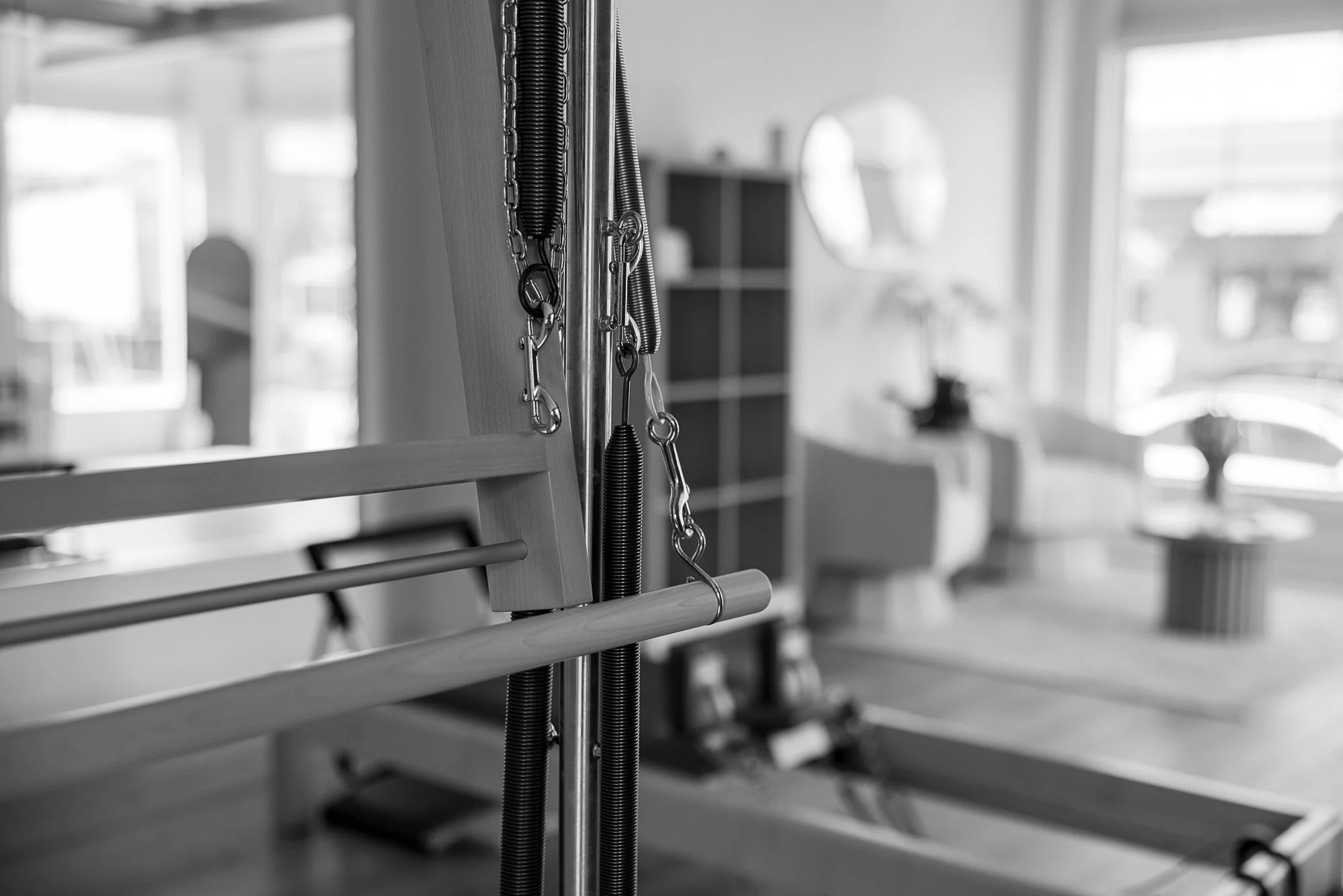 Close-up of a Pilates reformer machine with resistance springs and metal chains, in a bright, modern fitness studio.