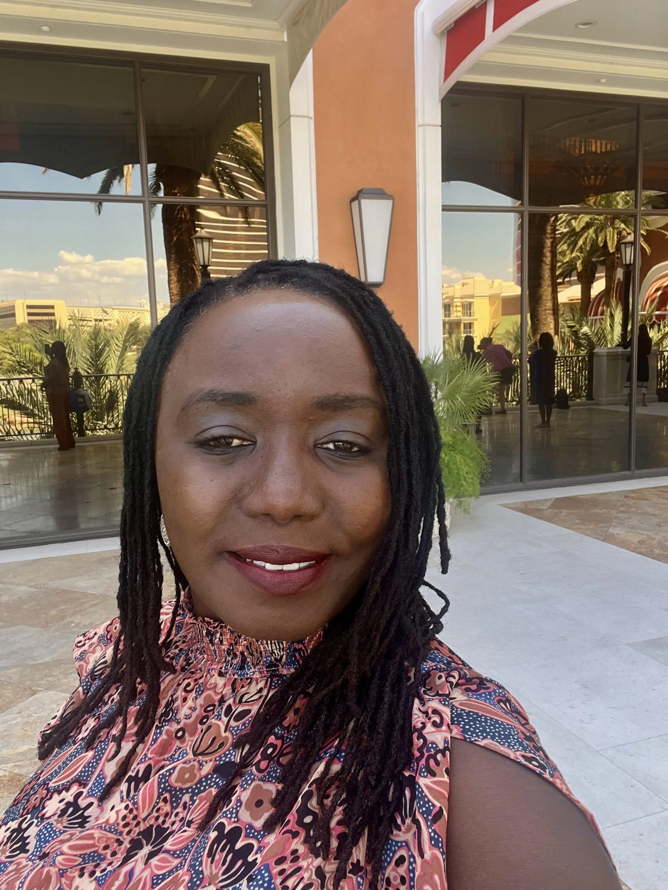 A woman with dark skin and shoulder-length dreadlocks taking a selfie outdoors in front of a building with large glass windows and tropical plants.