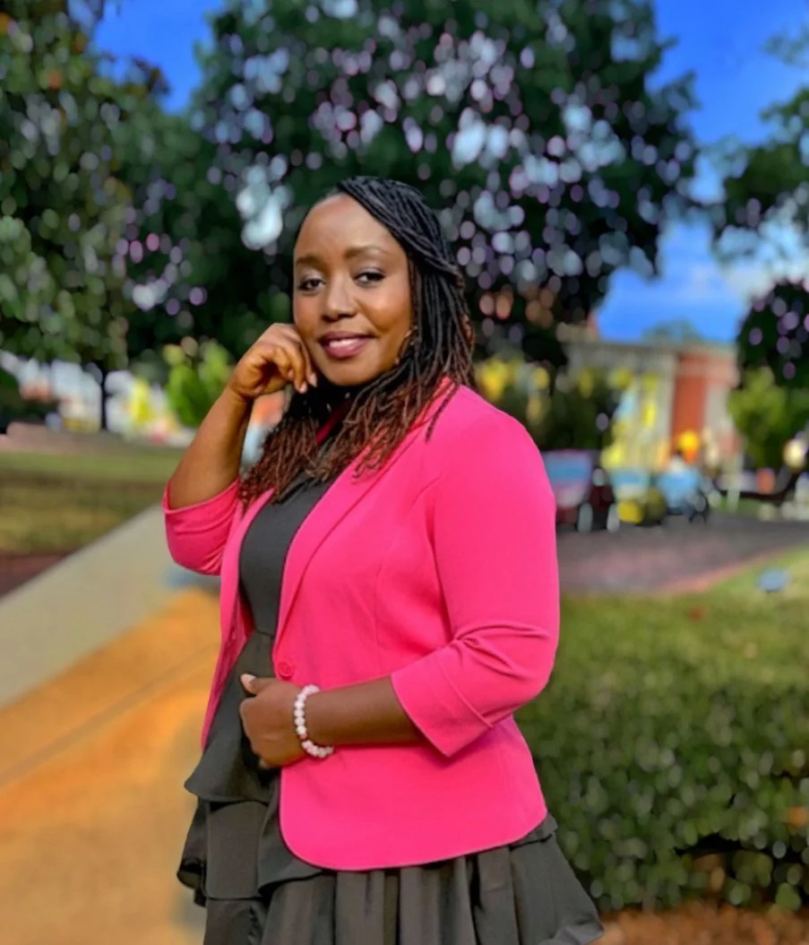 A woman wearing a bright pink blazer over a black top and a black skirt, standing outdoors with a blurred background of trees, cars, and a pathway.