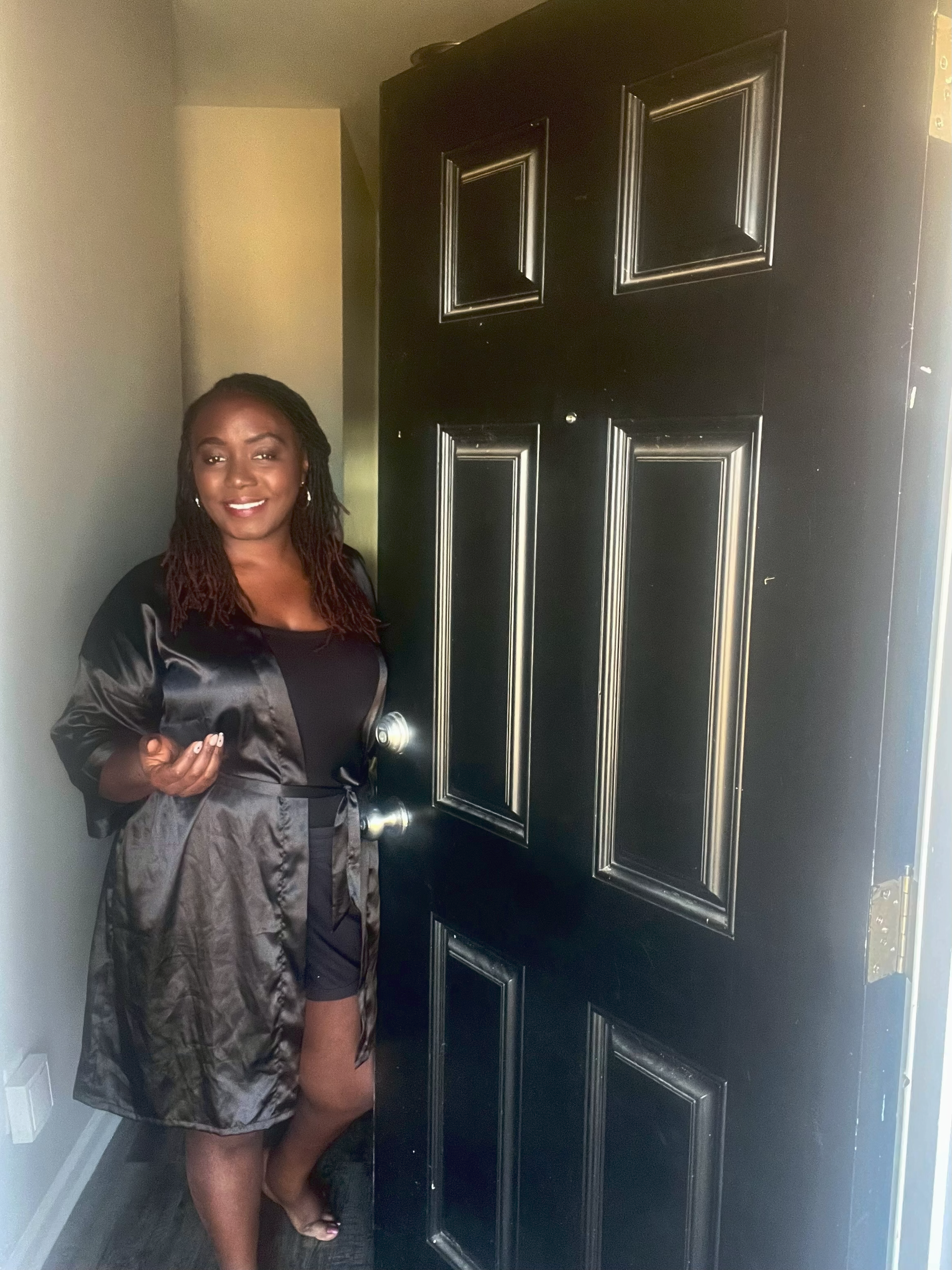 A woman standing in a doorway inside a house, smiling at the camera, wearing a black top and a dark gray satin robe or jacket.