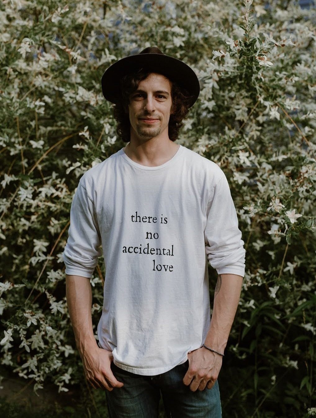 A young man with curly hair and a goatee, wearing a black hat and a white long-sleeve shirt with the words 'there is no accidental love' printed on it, standing in front of a bush with white flowers.
