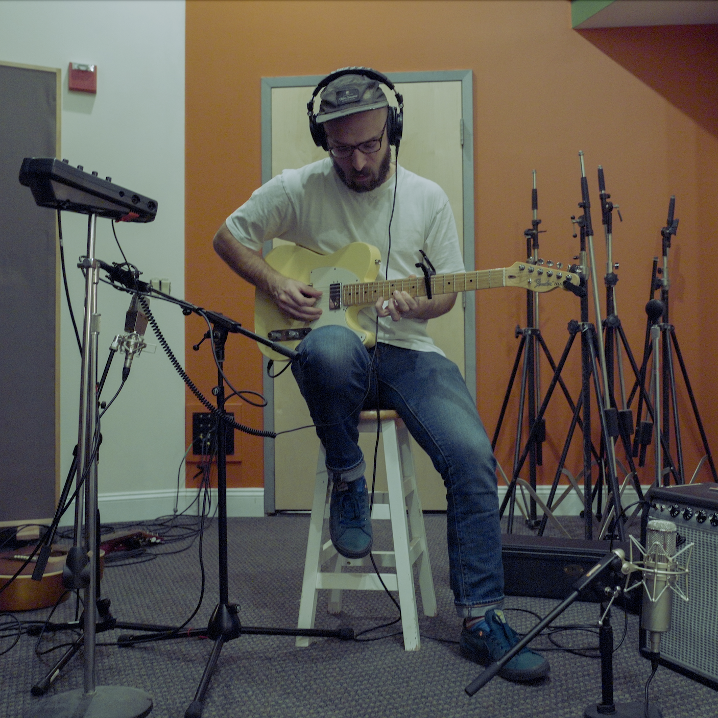 A man wearing headphones and glasses playing an electric guitar in a recording studio surrounded by microphones and music equipment.