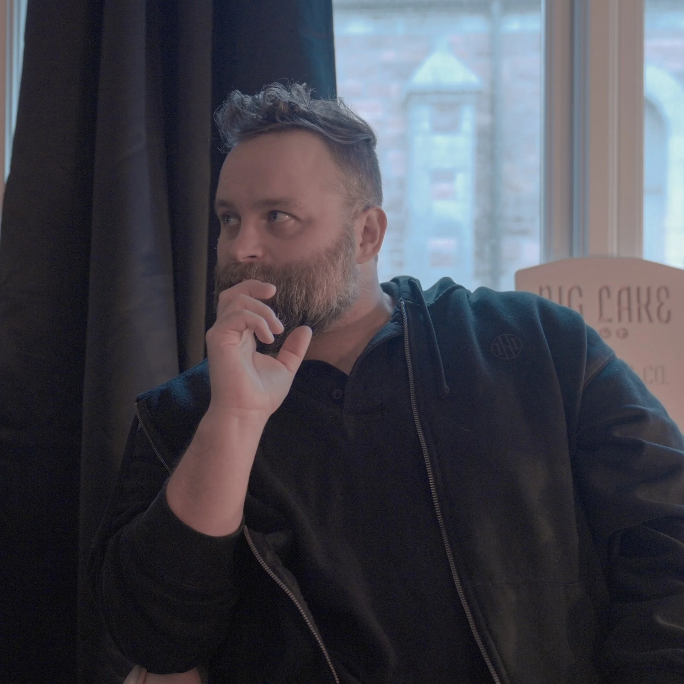A man with a beard and curly hair sitting indoors near a window, resting his chin on his hand and looking to the side.