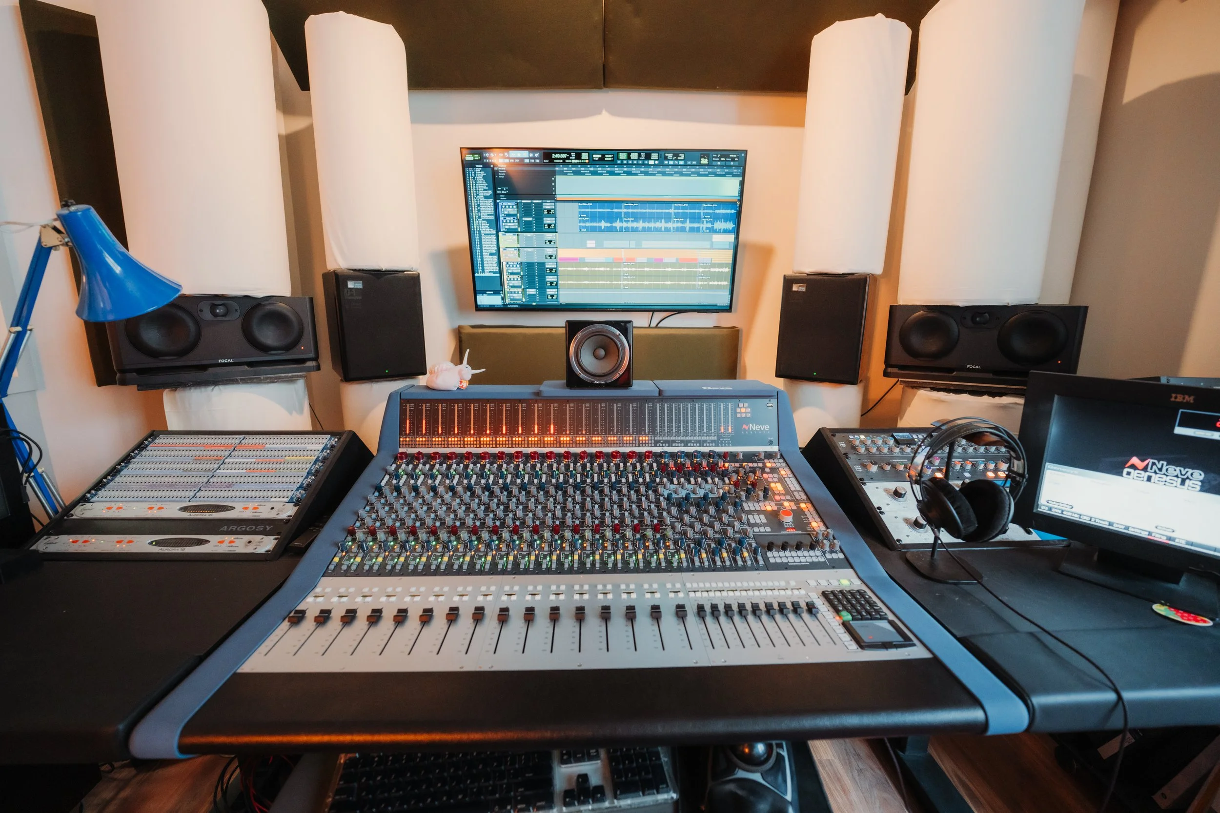 Music production studio with a large mixing console, audio equipment, speakers, a computer monitor, and headphones.