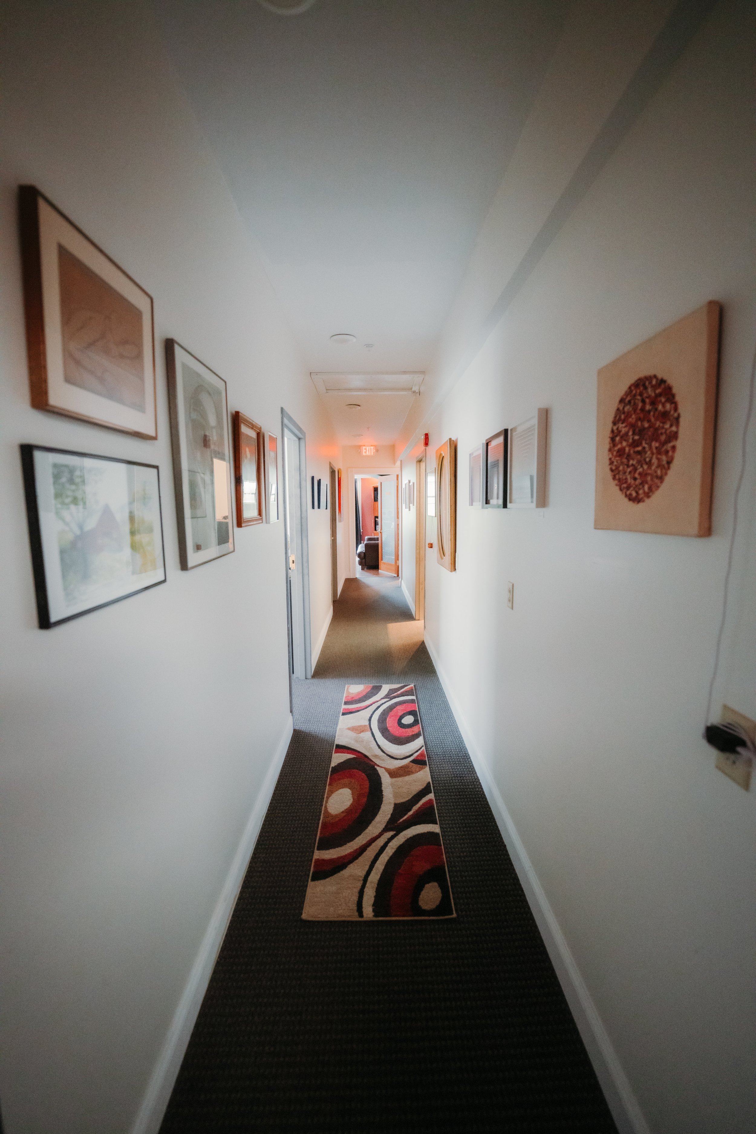 Long hallway with framed artwork on white walls, a colorful patterned rug on dark carpet, and a door at the end. Light gray ceiling with a few ceiling fixtures.