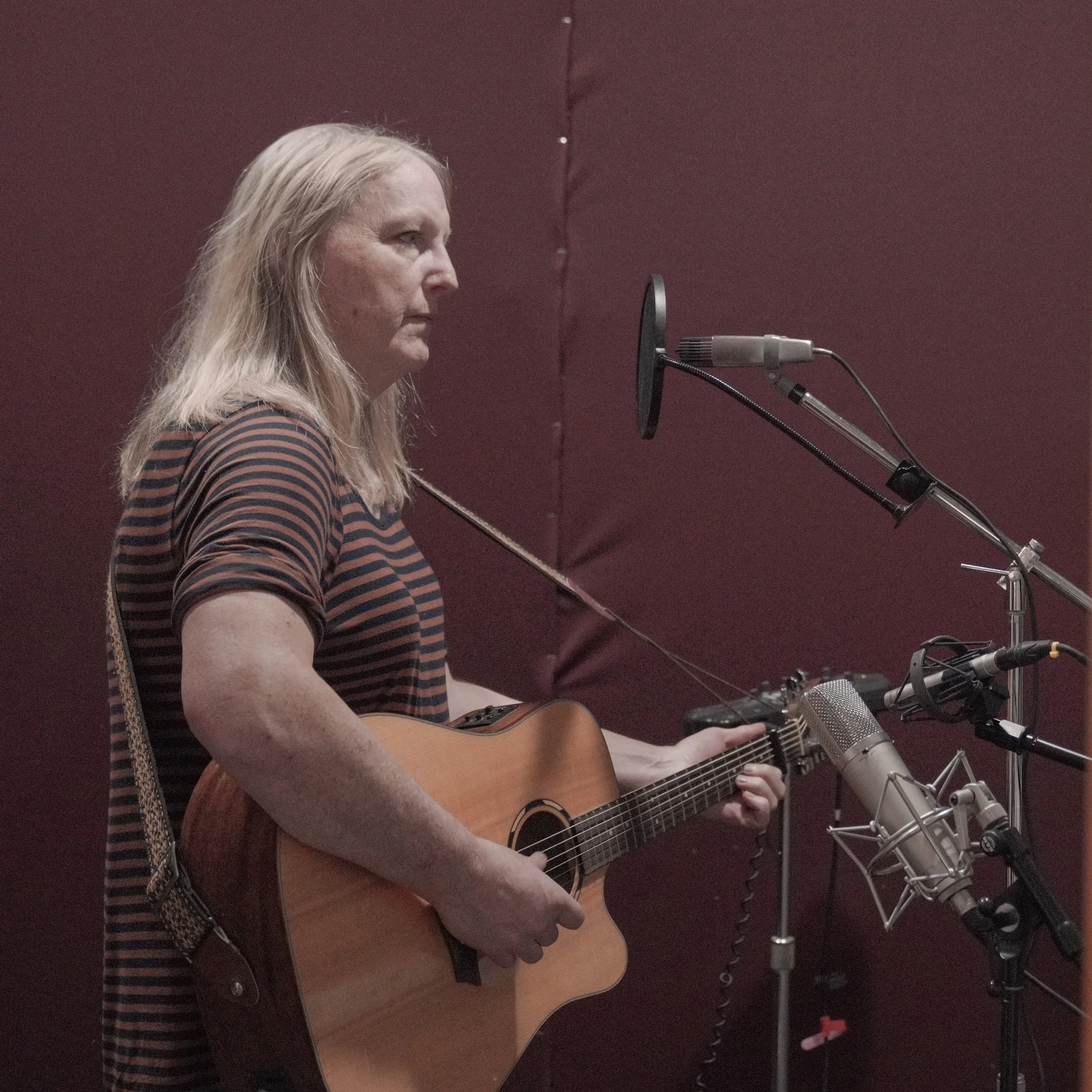 A woman with long blonde hair playing an acoustic guitar in a recording studio with a maroon-colored wall and professional microphones.