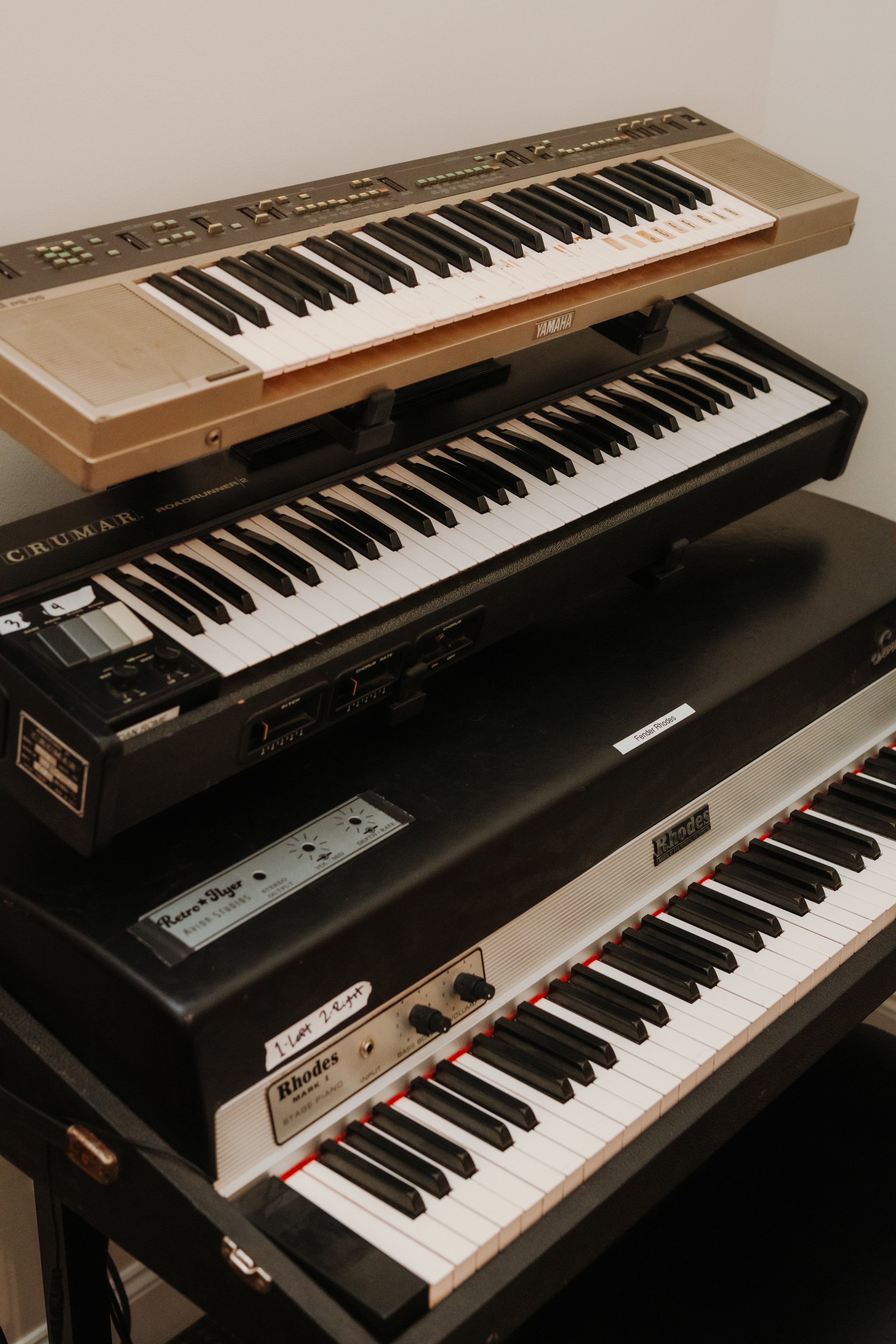 Three electronic keyboards stacked vertically on a black stand, with the top one being a Yamaha, the middle one a Crumar Roadrunner, and the bottom one a Rhodes electric piano.