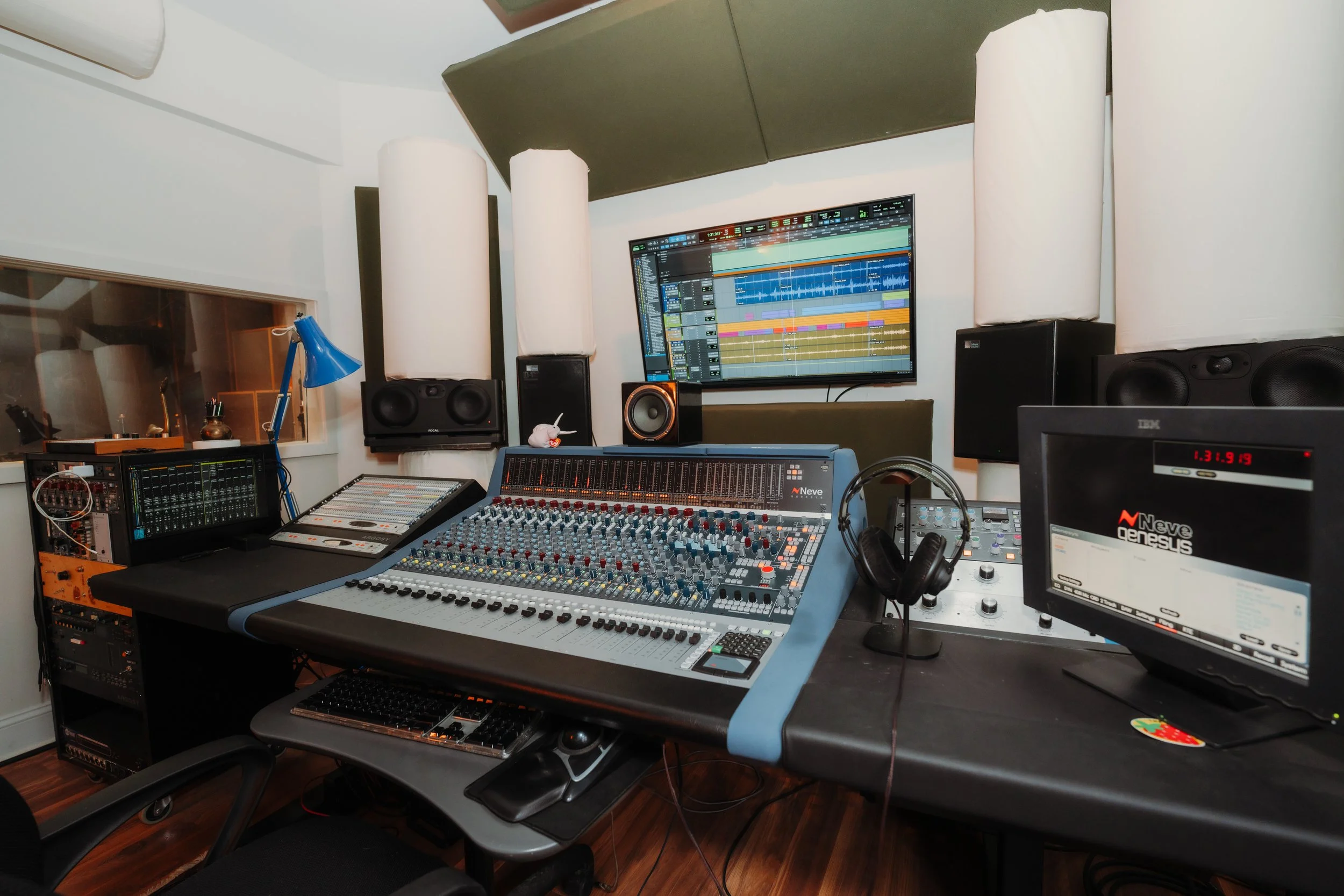 Music production studio with mixing console, computer monitors displaying digital audio workstations, studio speakers, and various audio equipment.