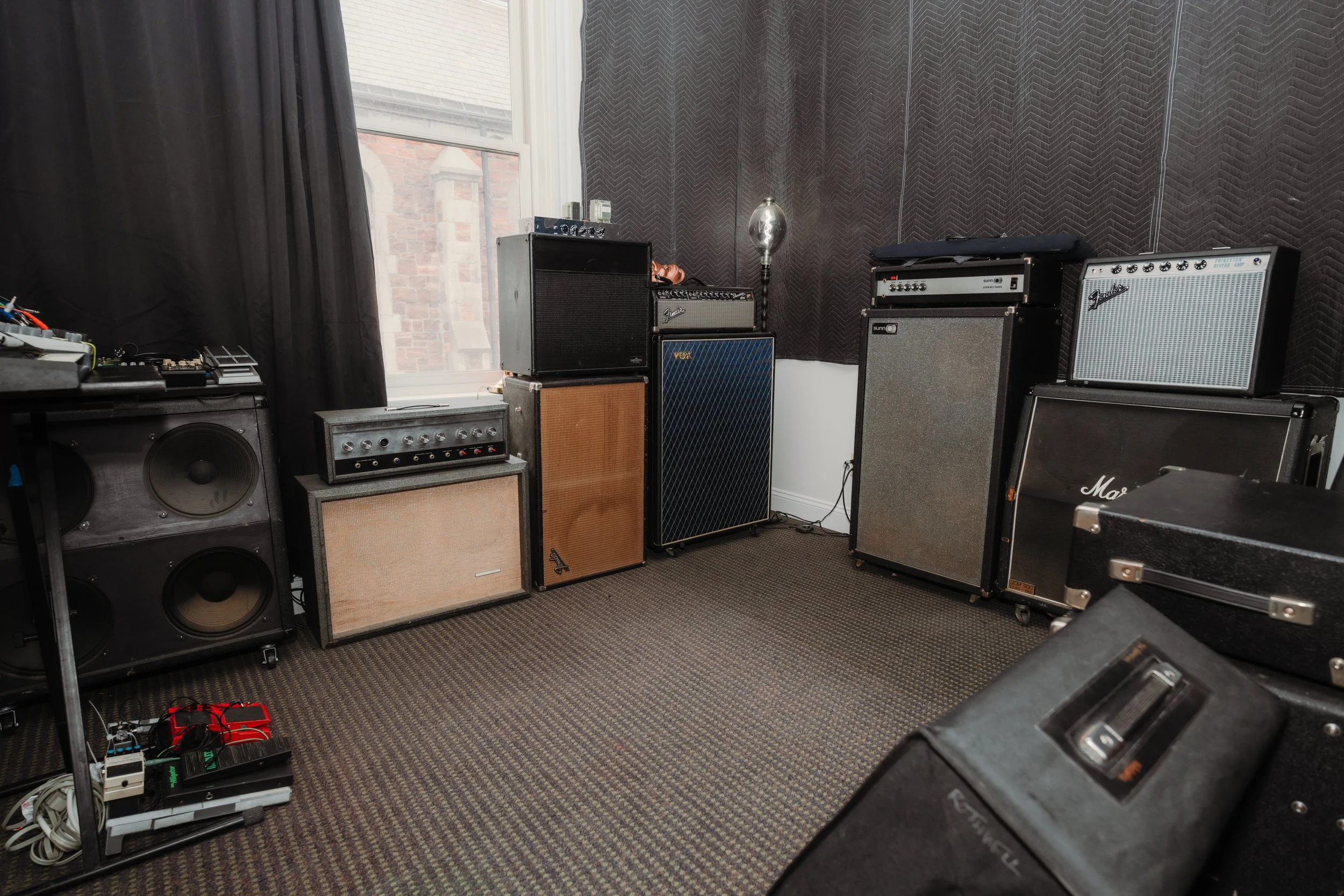 Music rehearsal room with various guitar amplifiers, speakers, and equipment stacked against dark wall panels near a window.