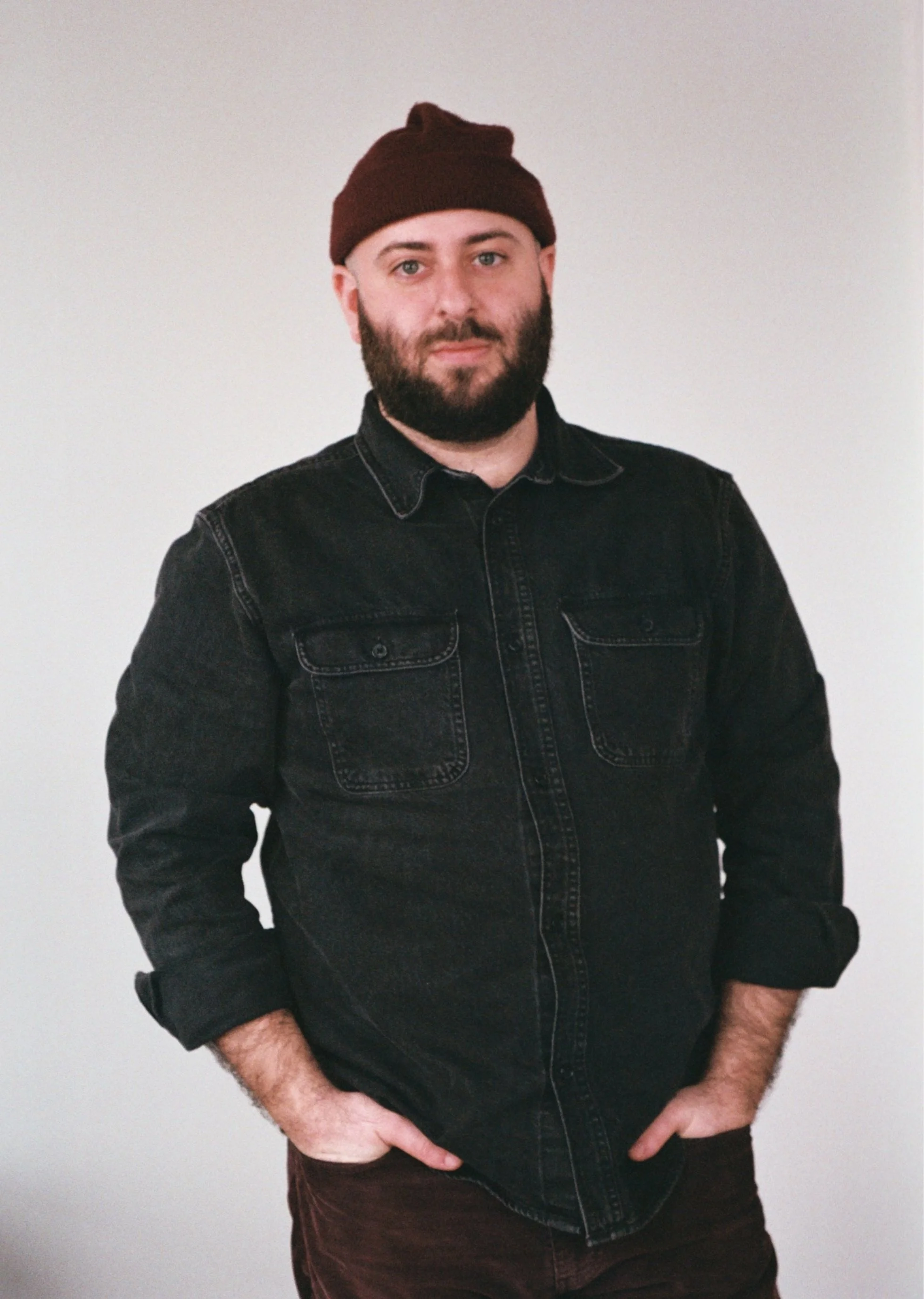 A man with a beard wearing a black denim jacket and a brown beanie, standing against a plain white background.