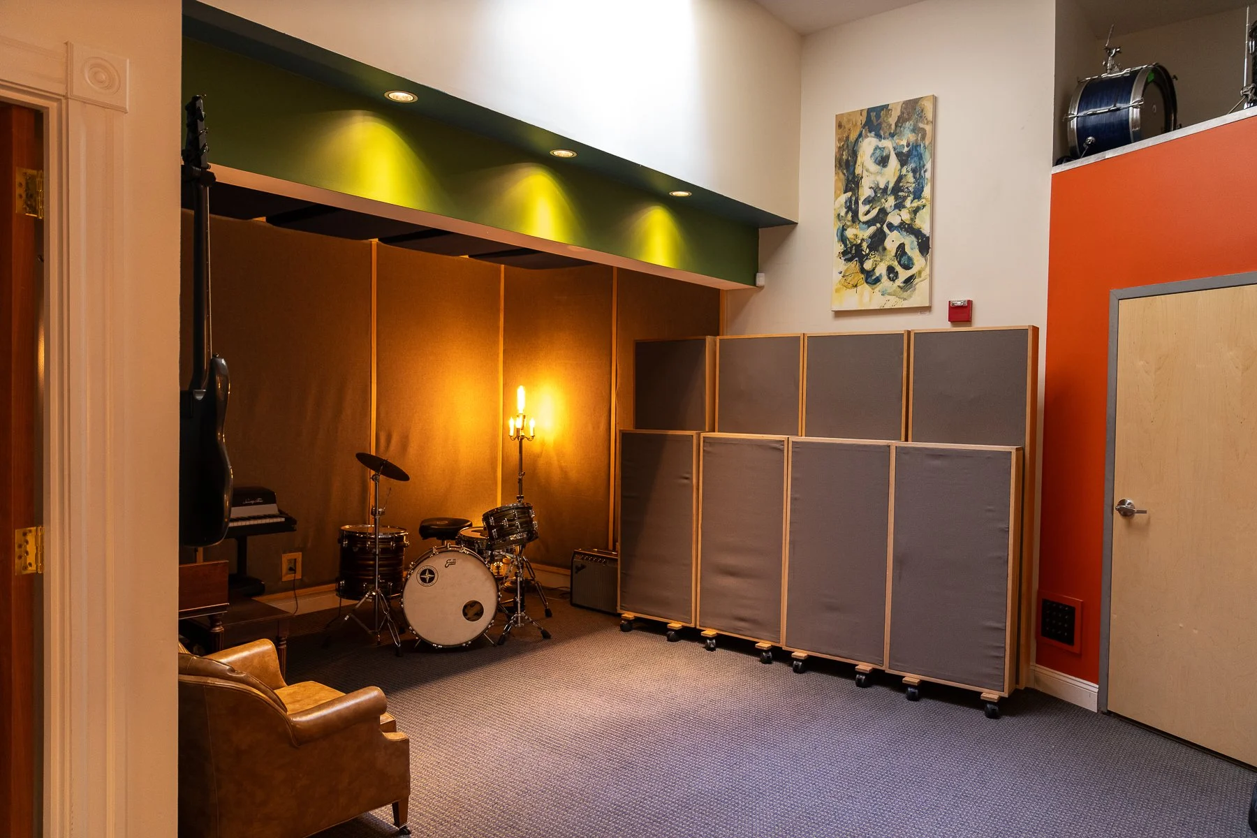 Music rehearsal room with drums, a guitar hanging on the wall, an armchair, acoustic panels, and a decorative wall lamp.