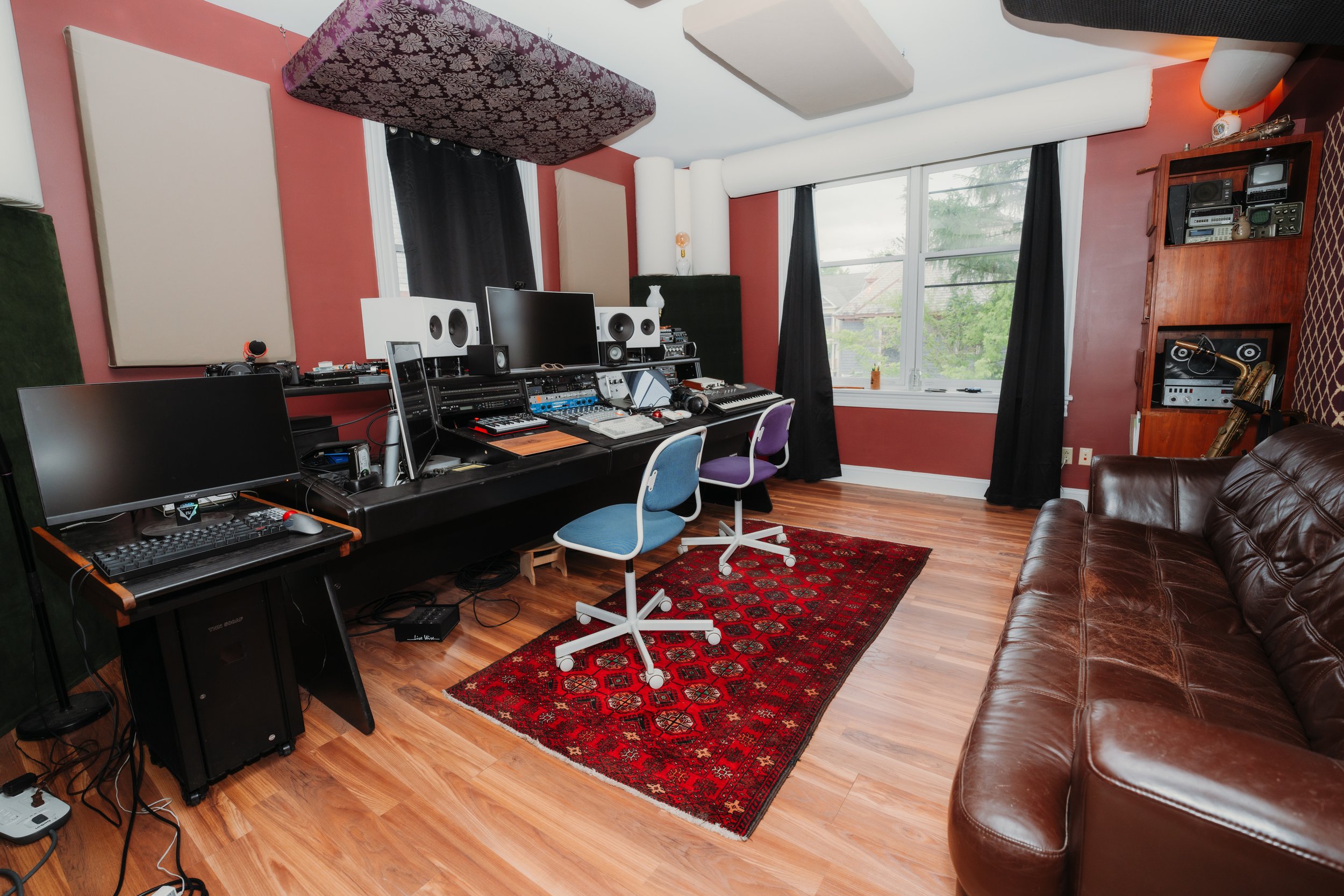 A music studio with a red wall, black curtains, and wood flooring. Contains multiple computers, speakers, and audio equipment on a long black desk. There is a brown leather couch on the right and a red patterned rug in the center.