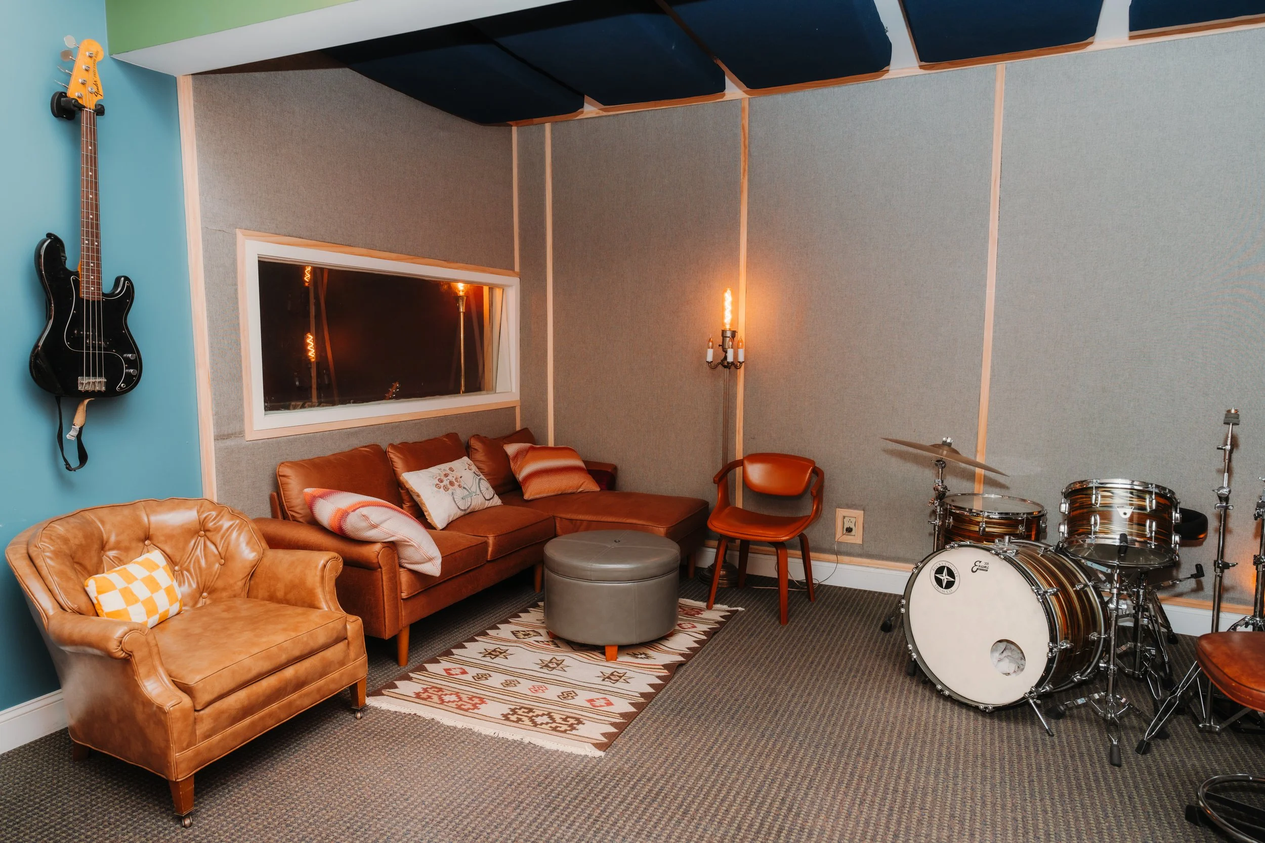 Music studio with vintage furniture, a guitar on the wall, a drum set, and a window reflecting light.