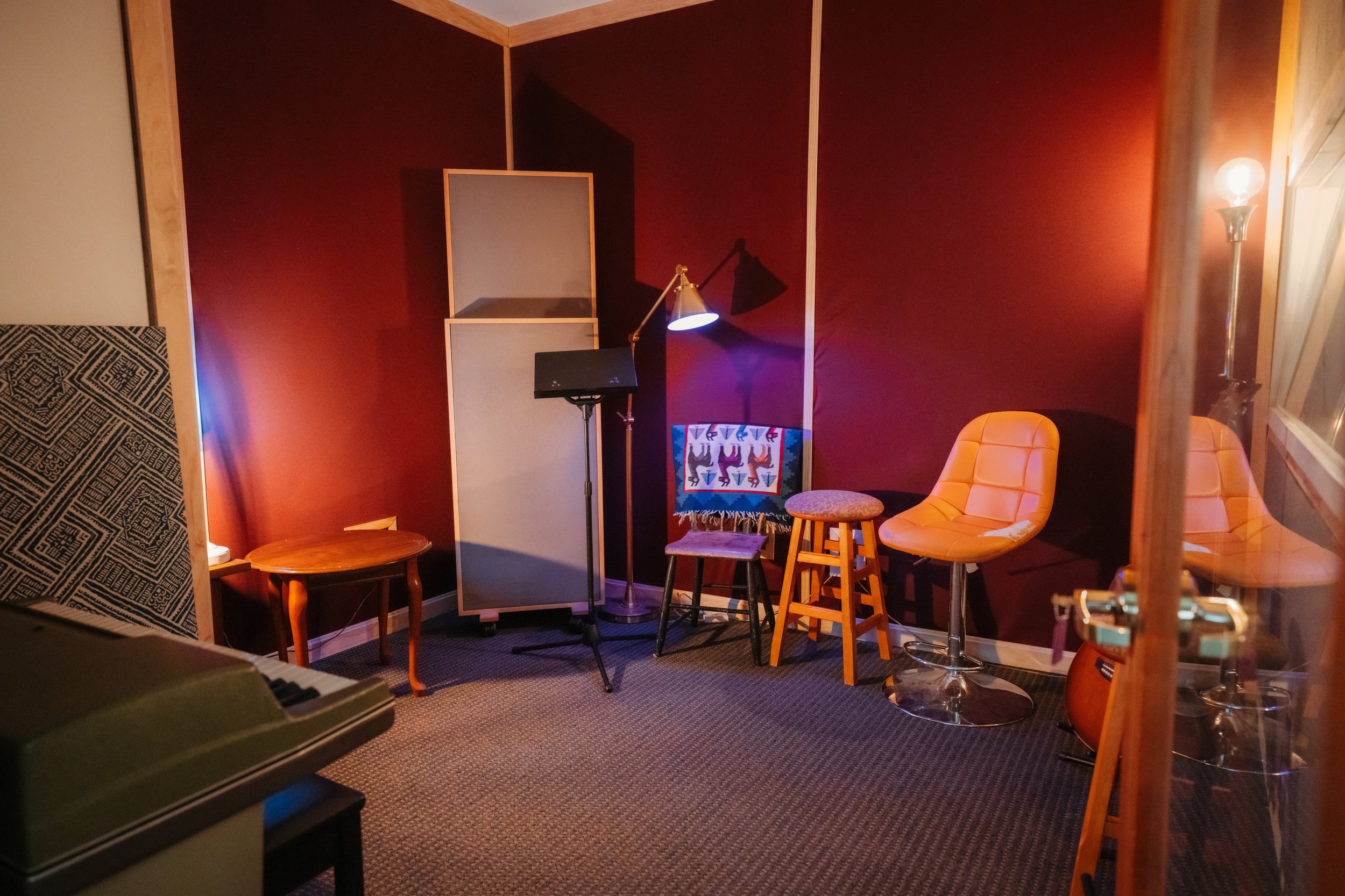 A small music studio with a red wall, featuring a vintage microphone stand, a music stand, two bar stools, a small wooden table, a vintage papasan chair, a guitar, and a standing floor lamp.