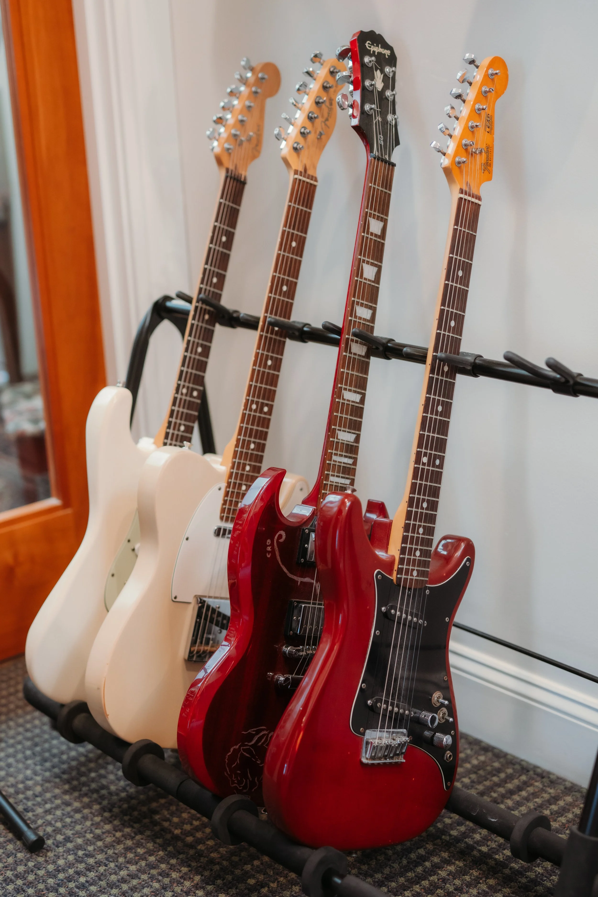 Four electric guitars on a wall-mounted rack, with two classic Fender Stratocasters and two other distinctive models, one black and one red, in a room with carpeted floor and a white walled background.