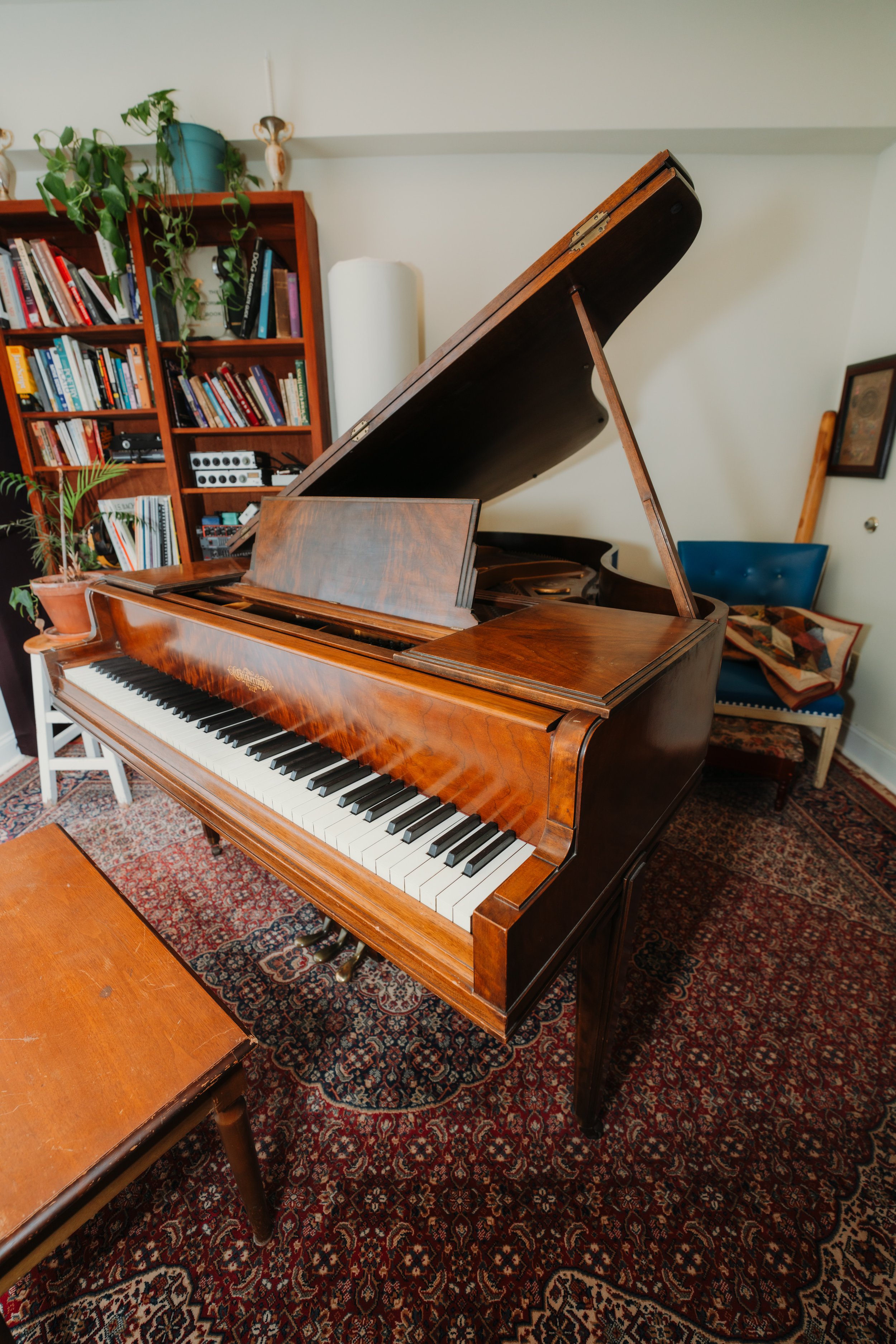 A wooden grand piano with an open lid, situated in a cozy room with a patterned rug, a wooden chair, a blue armchair with a quilt, and a bookcase filled with books and plants.