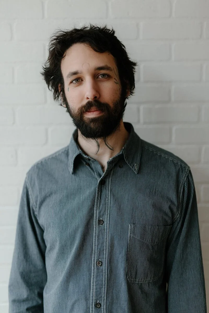 Portrait of a man with dark, wavy hair and a beard, wearing a gray striped button-up shirt, standing in front of a white brick wall.