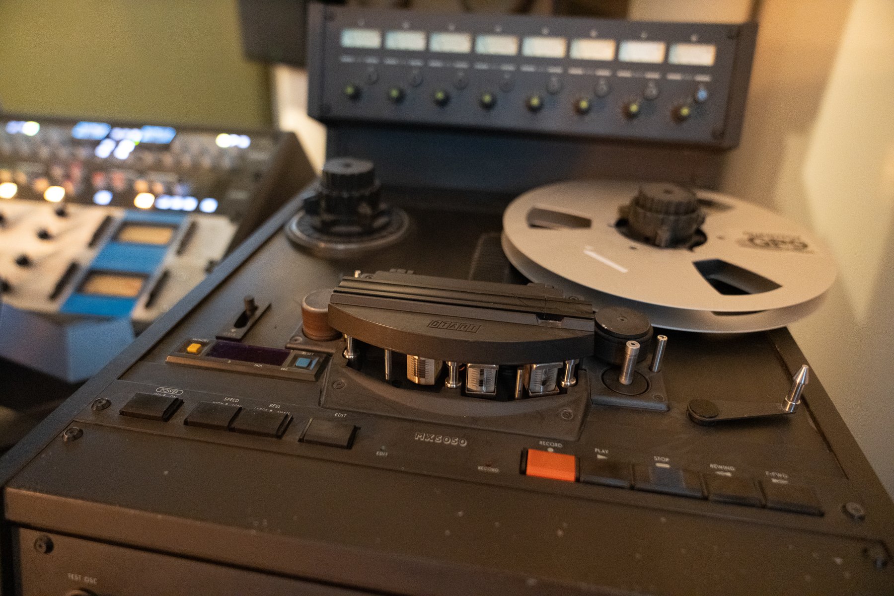 Close-up of a vintage reel-to-reel tape recorder with black and gray controls, a large reel of tape, and various knobs and buttons on a dark surface.