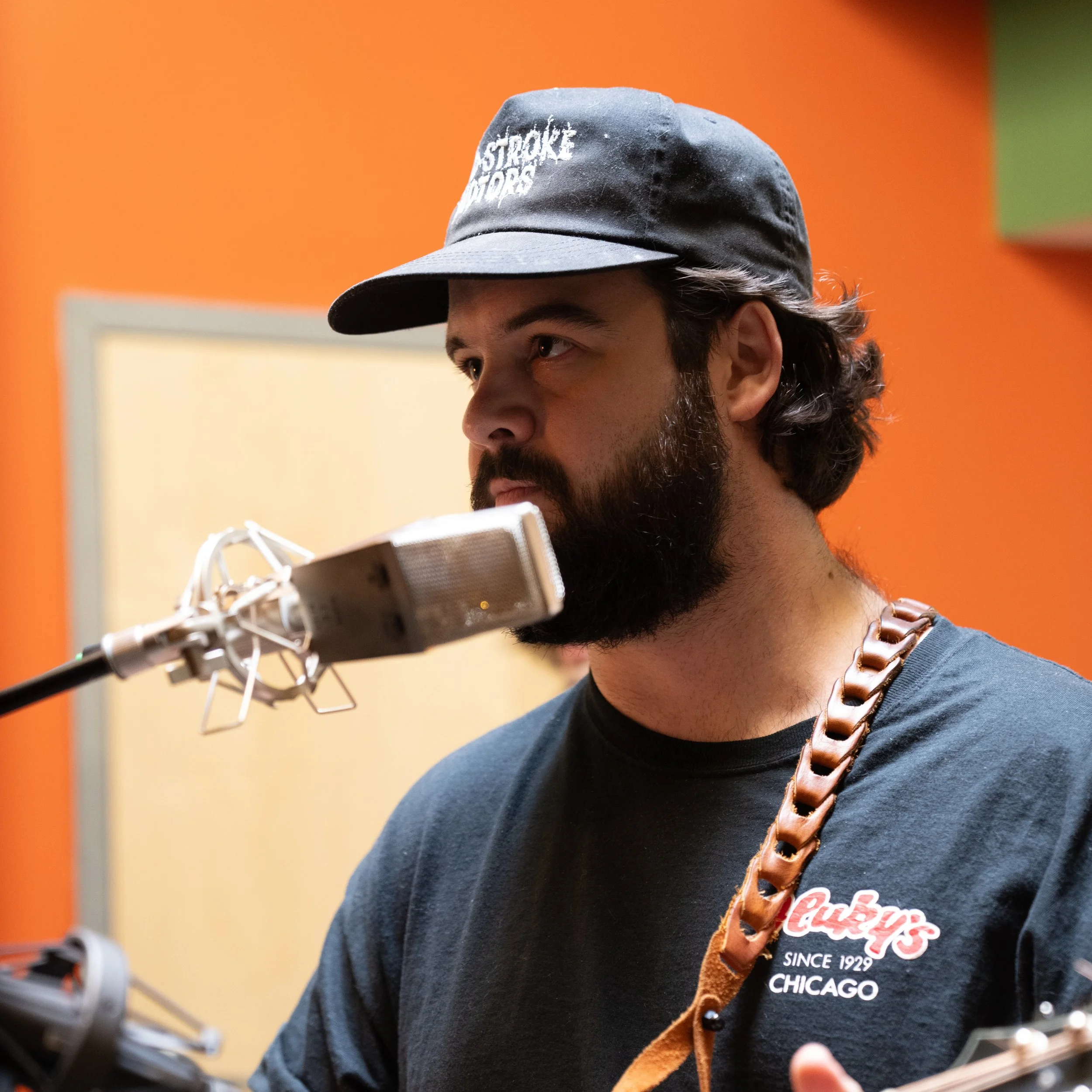 A man with a beard, wearing a baseball cap and a black t-shirt, singing into a microphone in a recording studio with orange and green walls.