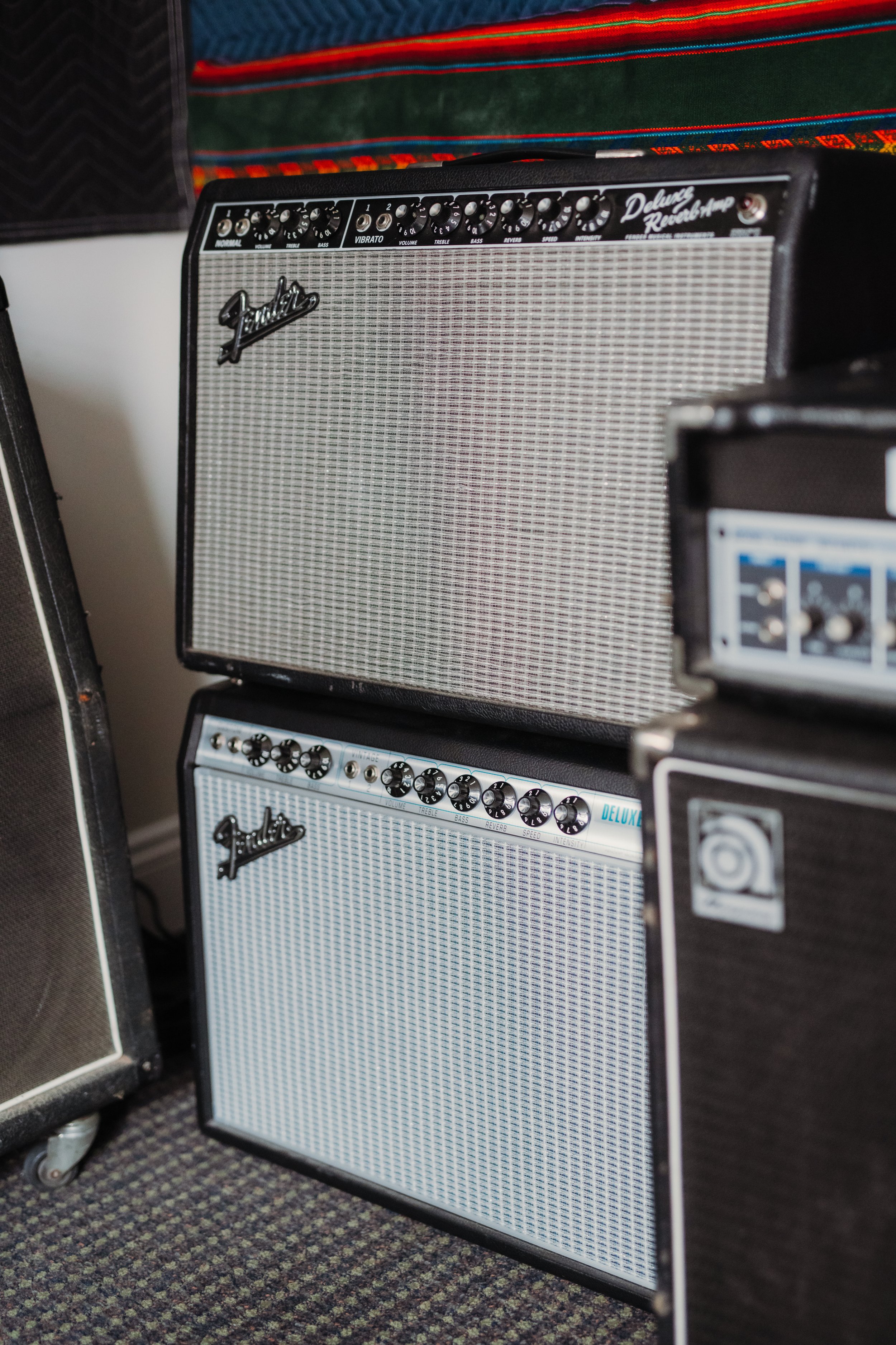 Two Fender guitar amplifiers stacked on top of each other, with various control knobs on top, in a room with framed textile art hanging on the wall.