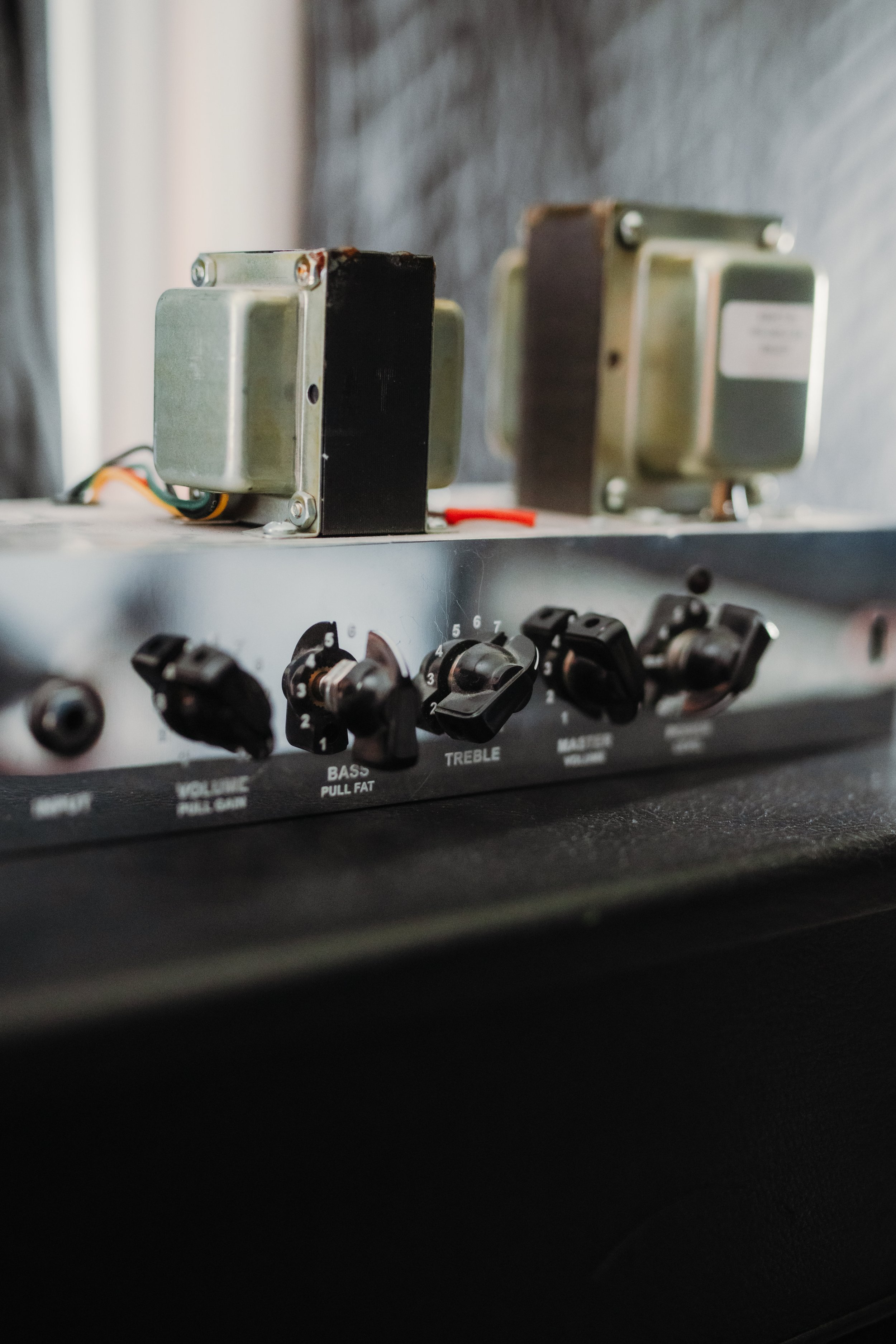 Close-up of a guitar amplifier control panel with knobs for volume, bass, treble, and master, and two transformers mounted on top.