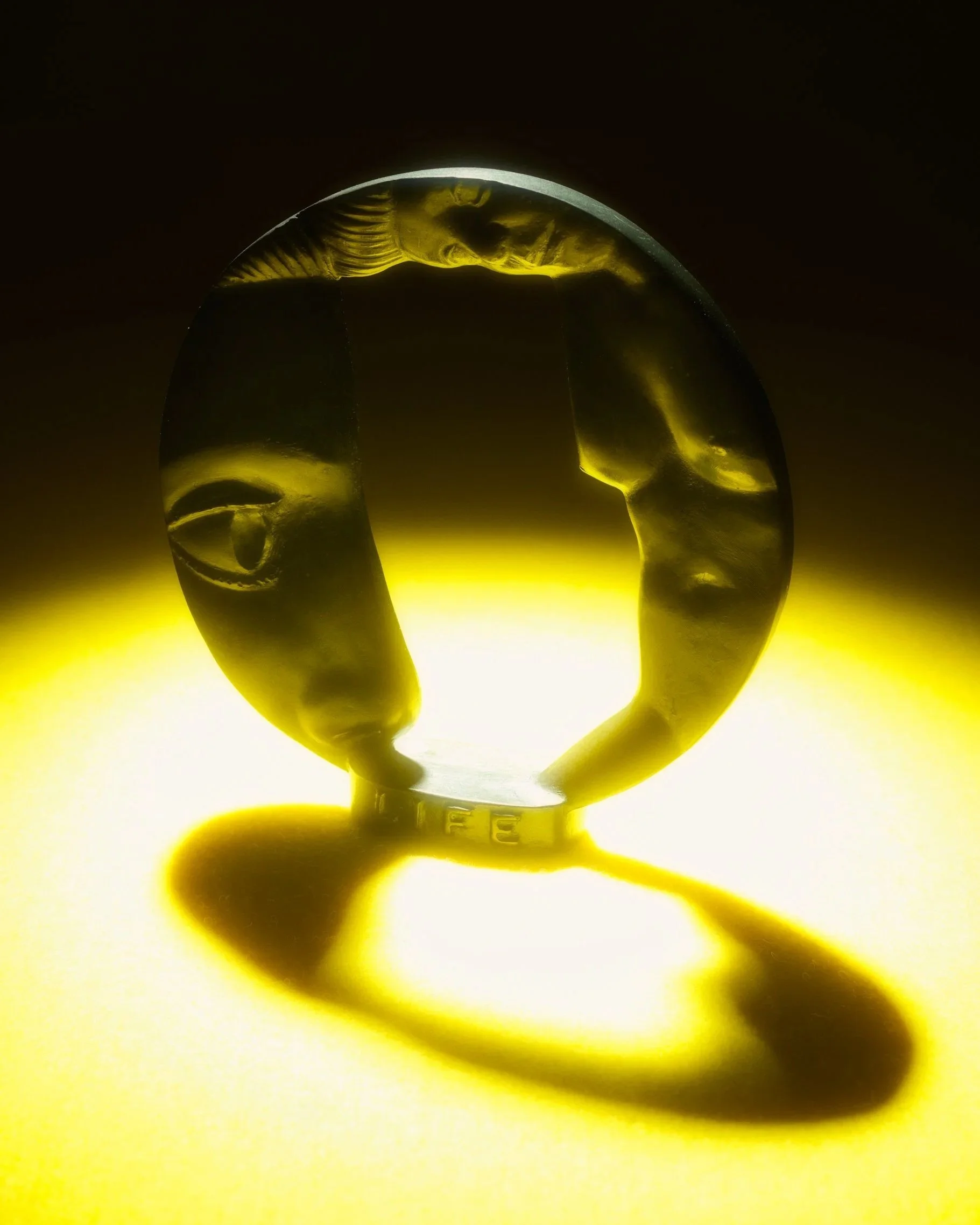 Still life photo of a circular bronze sculpture illuminated by a glowing light and casting a shadow in front.