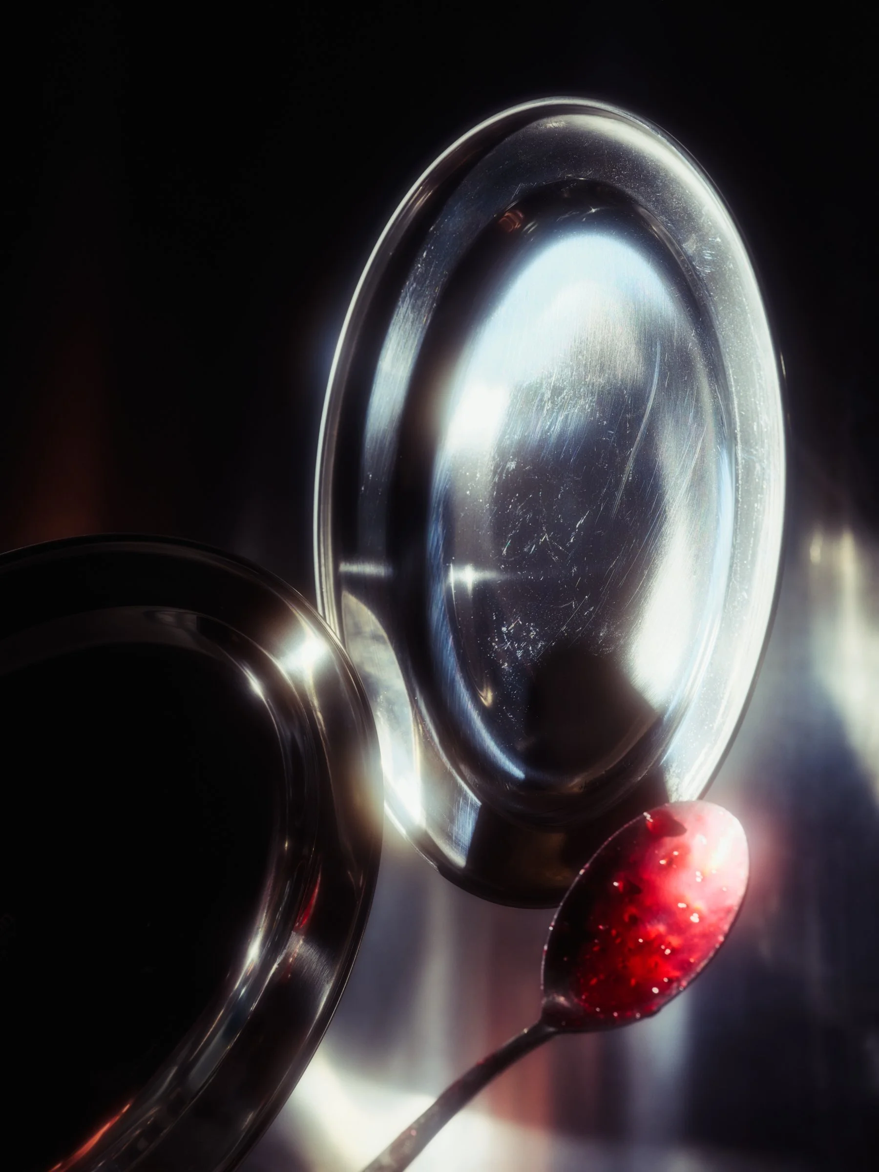 A still life image of silver plates with glowing highlights and a spoon covered in red jam