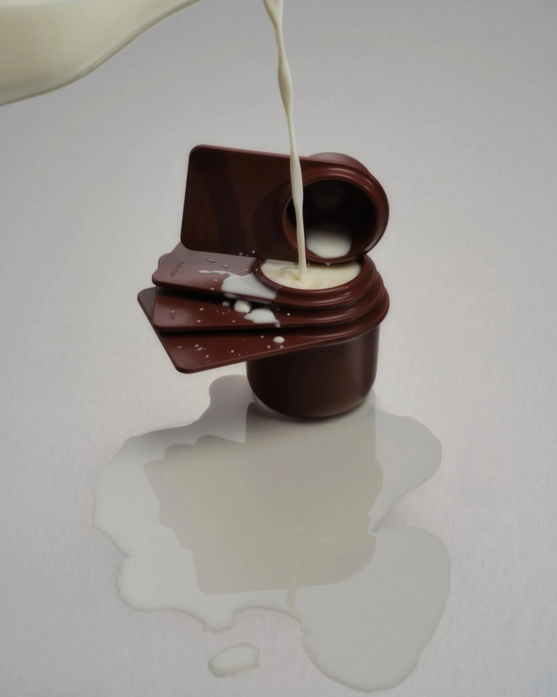 Photograph of milk being poured into 4 brown measuring cups overflowing onto a metal surface.