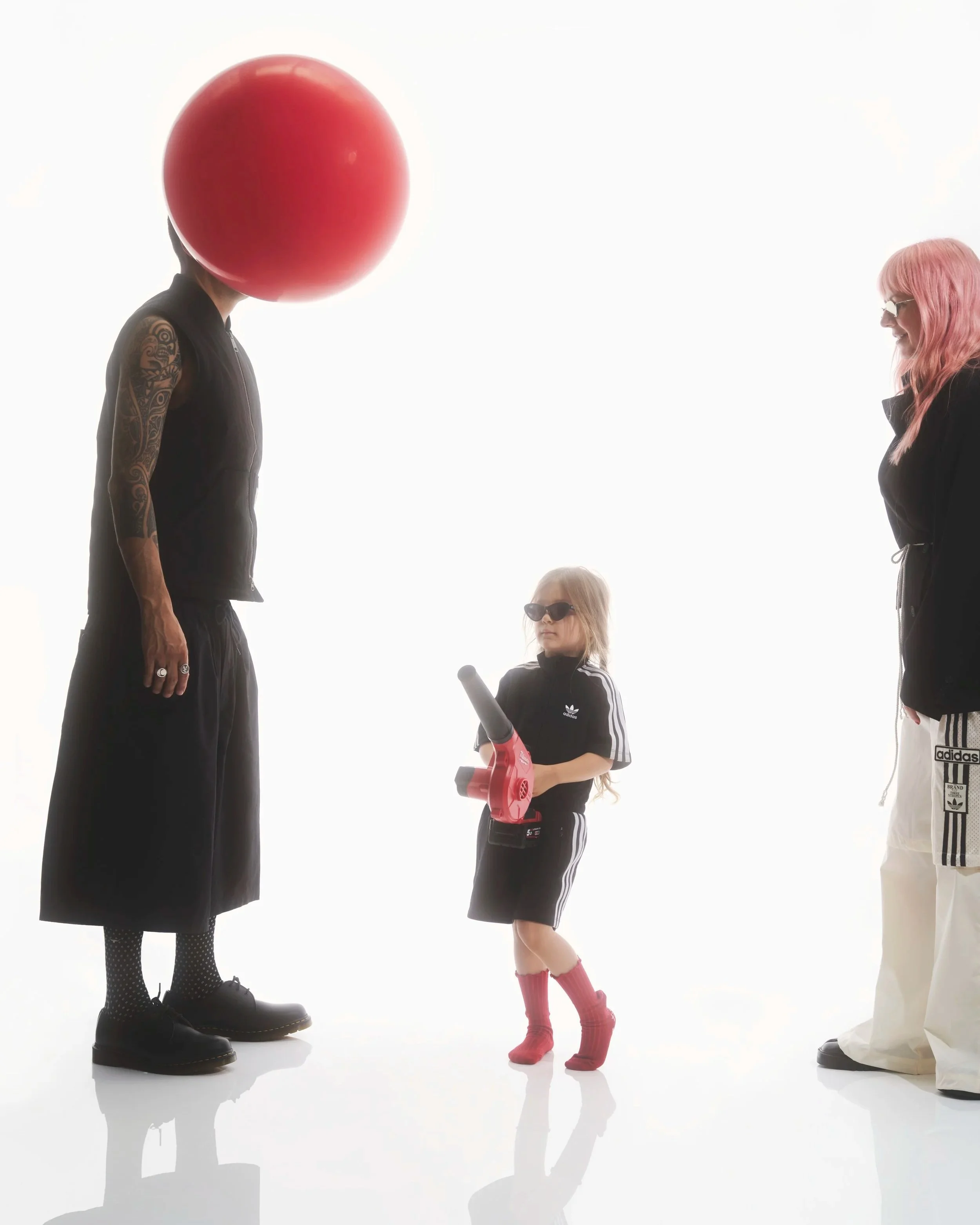 A portrait of three people on a white background. A small girl with a red leaf blower a man with a balloon over his head and a woman with pink hair.