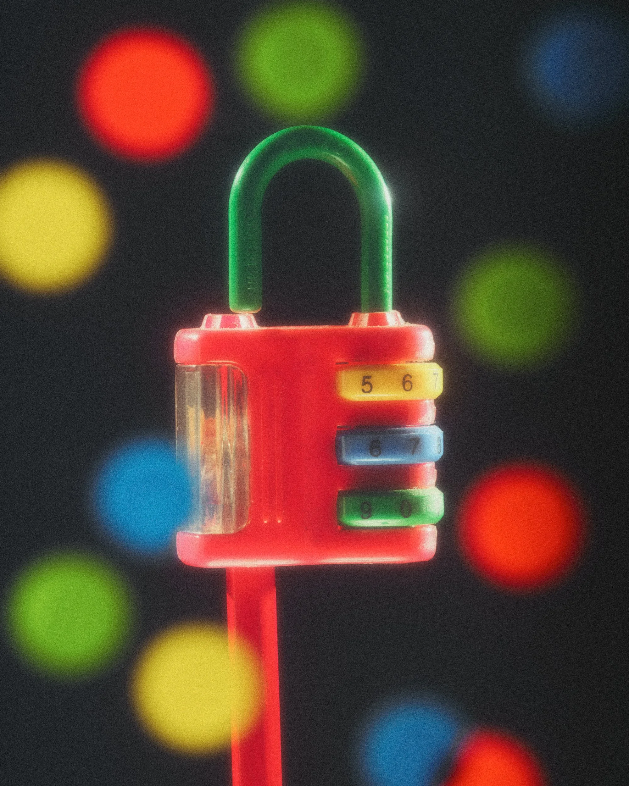 Still life photo by Sian Fay Kerr. A red pencil sharpener shaped like a lock with glowing highlights. Circular stickers in red, green, blue and yellow softly focused in the foreground.