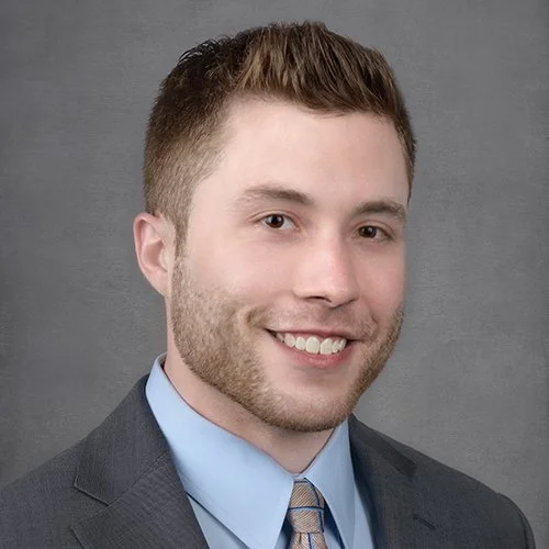Professional headshot of a young man with short brown hair, wearing a gray suit, light blue dress shirt, and light brown tie, smiling against a gray background.