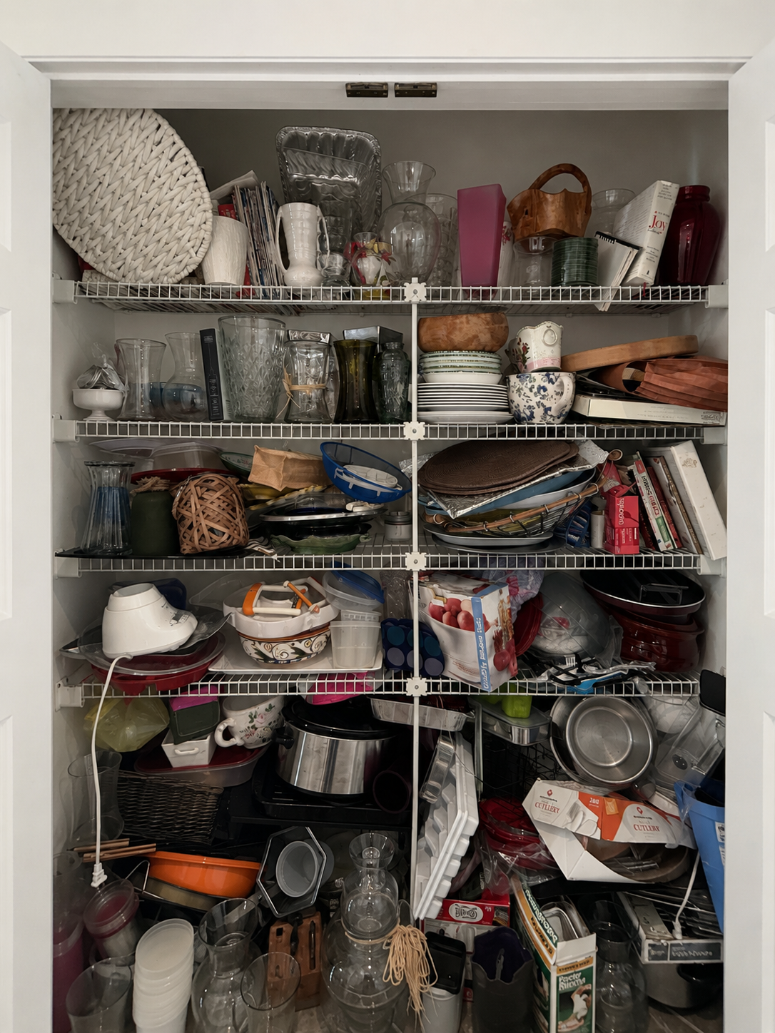 Overfilled kitchen storage closet with crowded shelves, stacked dishes, cookware, serving trays, and miscellaneous household items showing accumulation and home organization overwhelm.