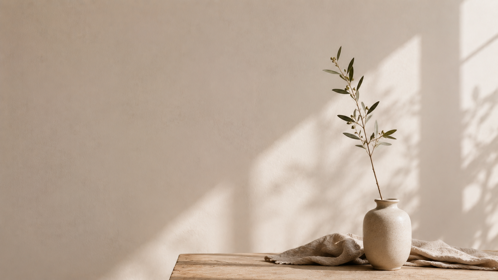 A beige ceramic vase with a single dried branch on a wooden surface, illuminated by natural sunlight, against a plain wall.