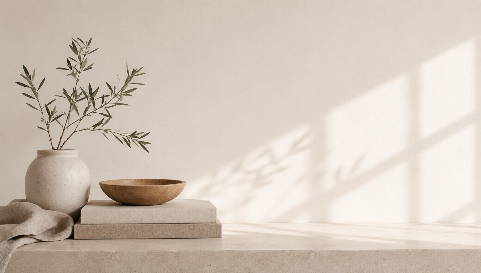 A beige ceramic vase with a simple, rounded shape, holding dried branches with small round seed pods, placed on a wooden surface with a plain, light-colored wall in the background.