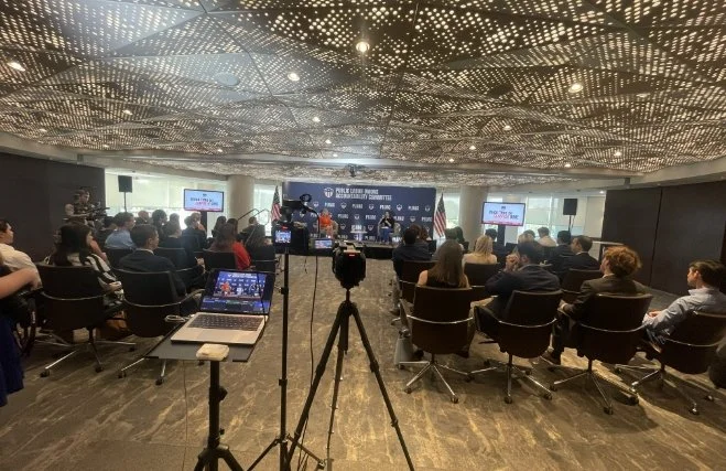 Conference room with a seated audience facing a stage with speakers. Cameras and laptops are set up in the foreground, and the ceiling has an intricate, illuminated pattern.