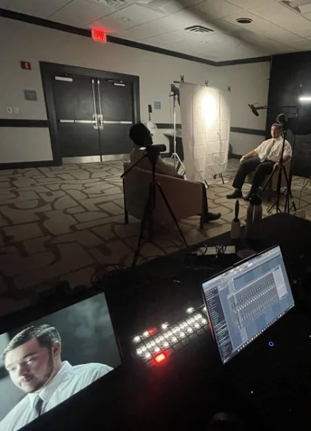 A behind-the-scenes look at a photo or video shoot setup. There are two people, one sitting in a chair with a microphone, and another standing or sitting near a white backdrop. Professional lighting and camera equipment are positioned around them. A monitor in the foreground displays a man’s face, and a laptop shows audio or video editing software.