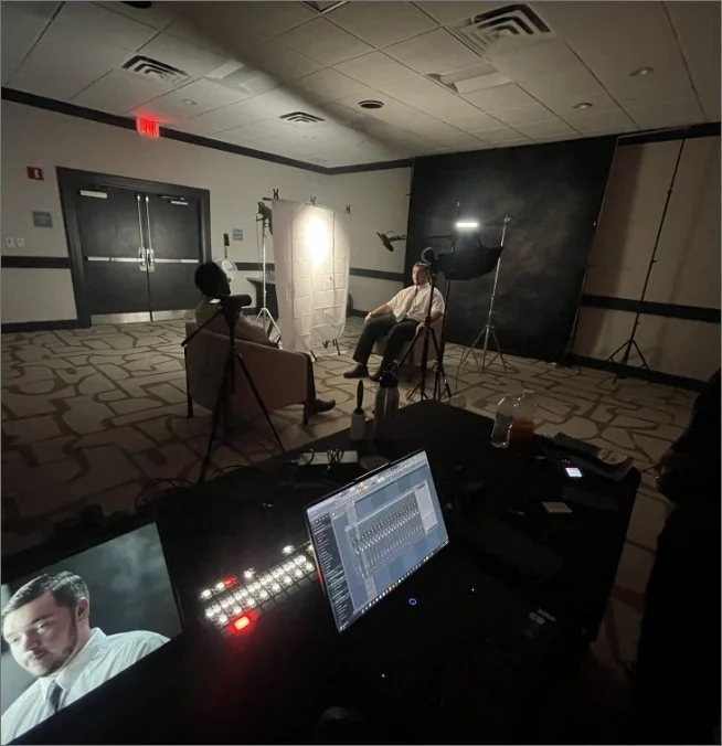 A behind-the-scenes look at a video or photography shoot in a dimly lit room. Two individuals are seated and talking, one with a microphone, with studio equipment and lighting setup around them. In the foreground, a monitor displays a close-up of a man with dark hair.