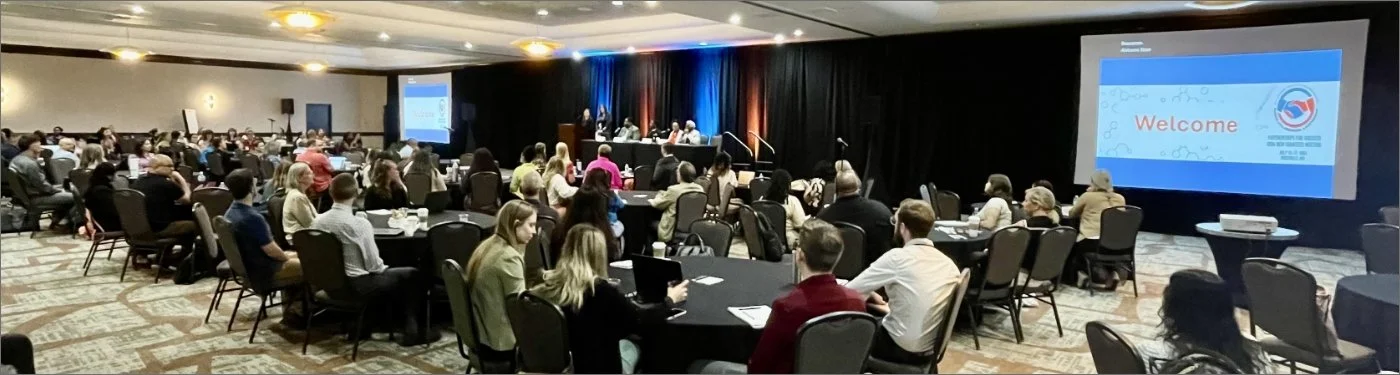 Conference room filled with attendees seated at round tables, facing a stage with a large screen displaying a welcome message, and panel discussion taking place at the front.