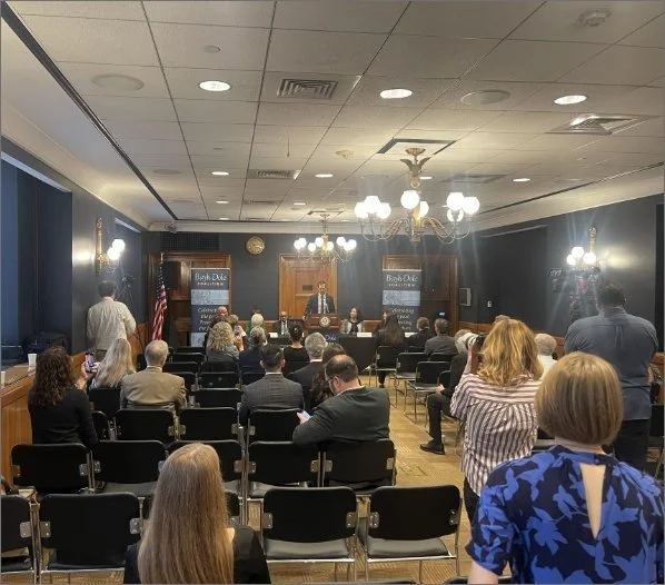 A meeting room with people attending a presentation or discussion. There is a speaker at a podium at the front, and banners with 'Bank Ozark' visible. The room has blue walls, chandeliers, and an American flag.