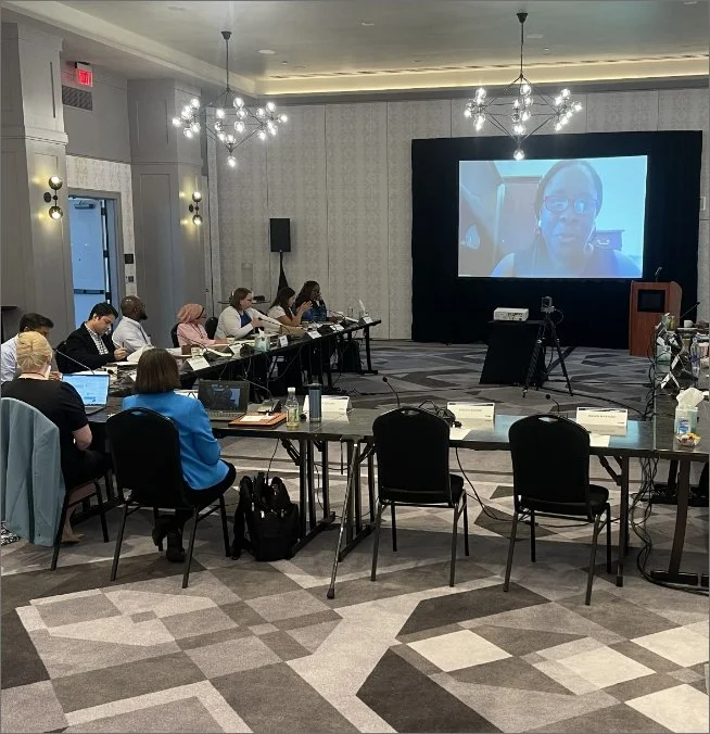 Conference room with attendees sitting at tables, participating in a video call projected on a large screen at the front of the room.