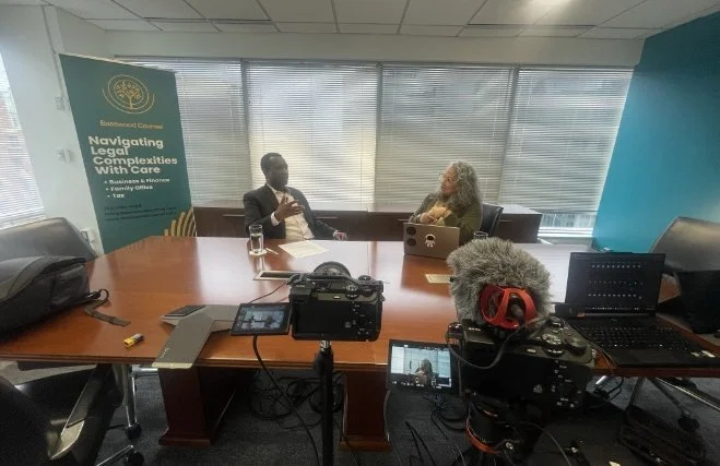 Two people sitting at a conference table in a video interview setup, with cameras and audio equipment recording. A banner on the left reads 'Navigating Legal Complexities with Care' and the room has large windows with blinds.